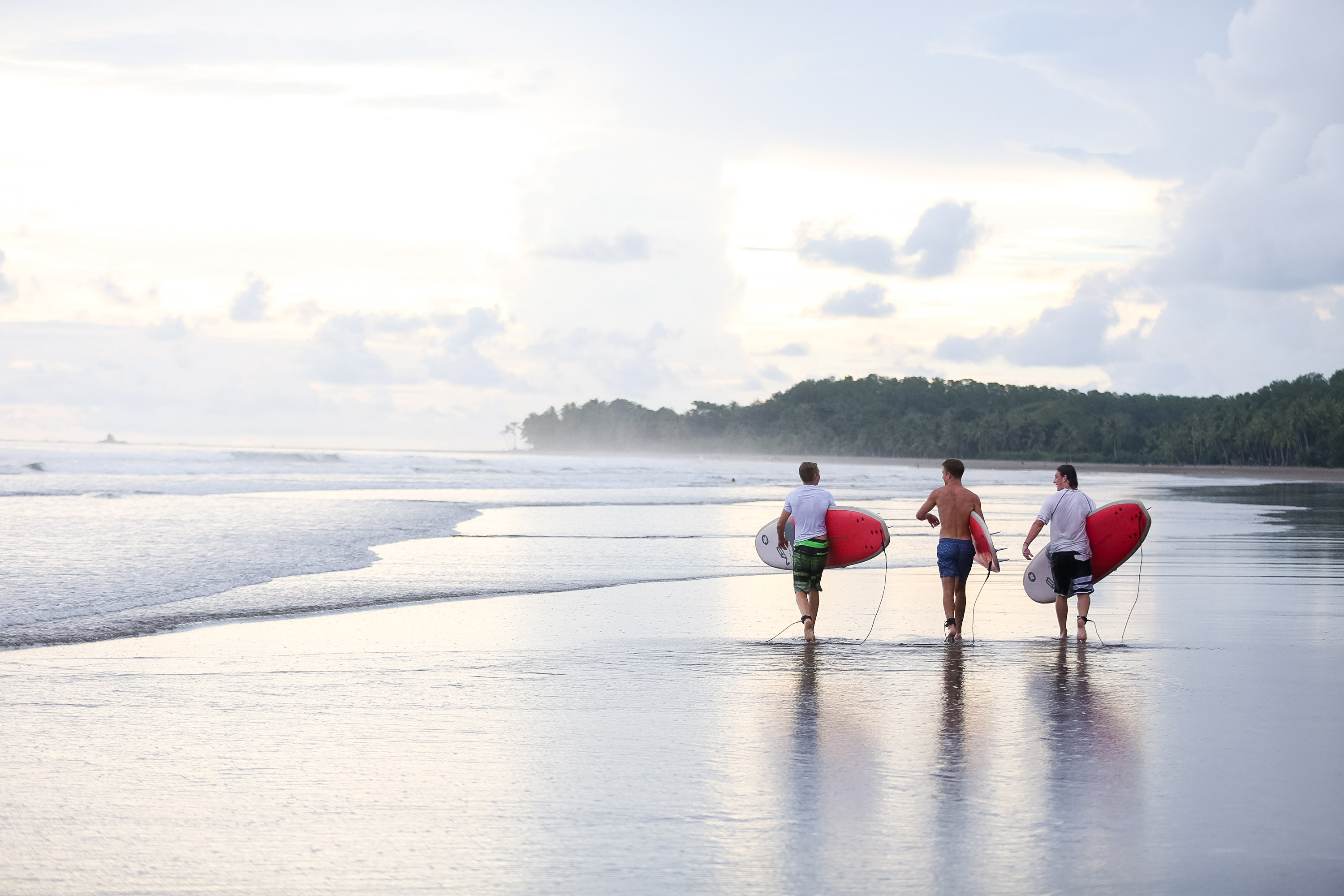 Students carry surfboards across a wet beach at dusk