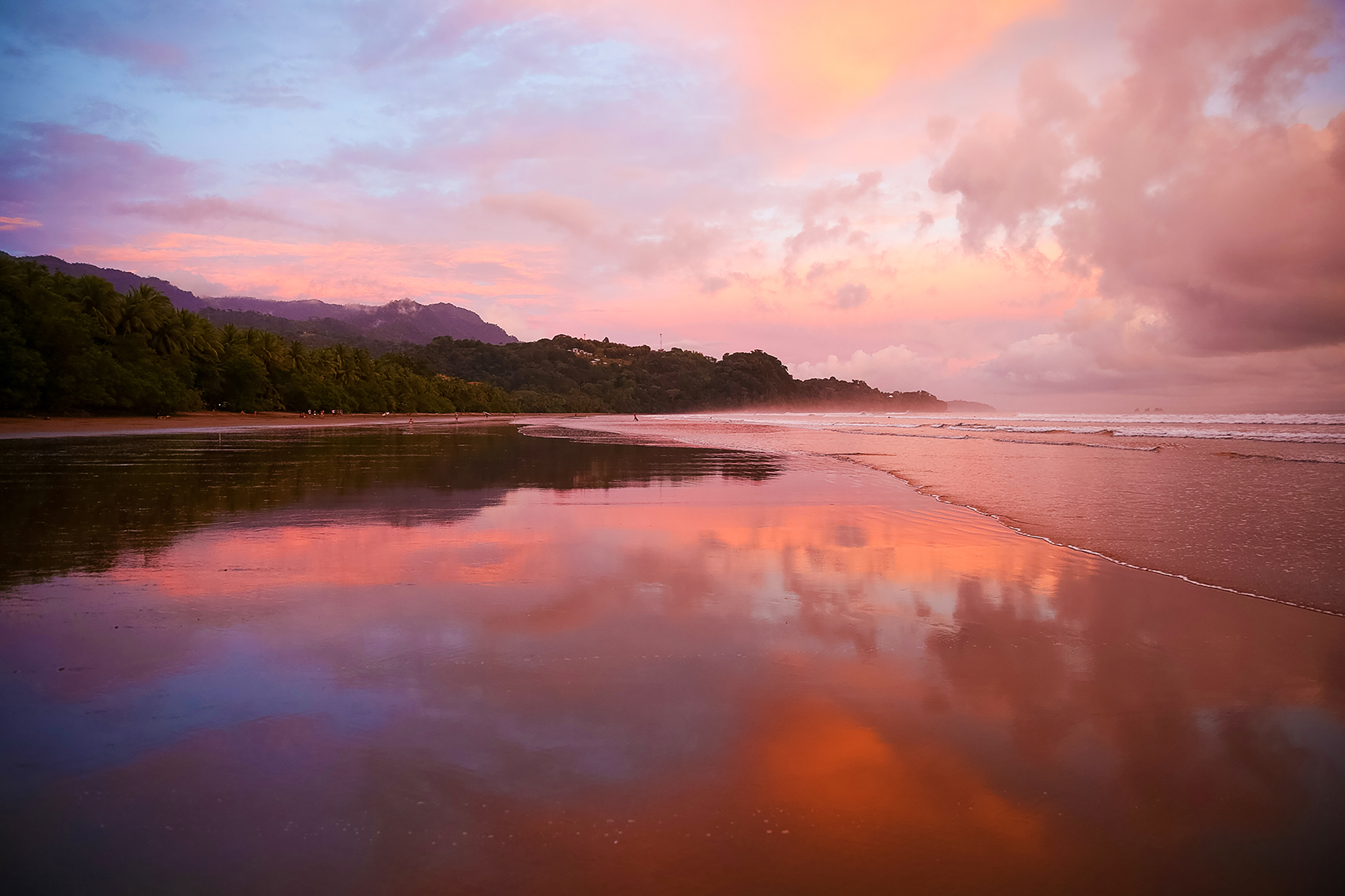 Wet sand mirrors a pink and orange sunset sky along a jungle-lined Costa Rica beach