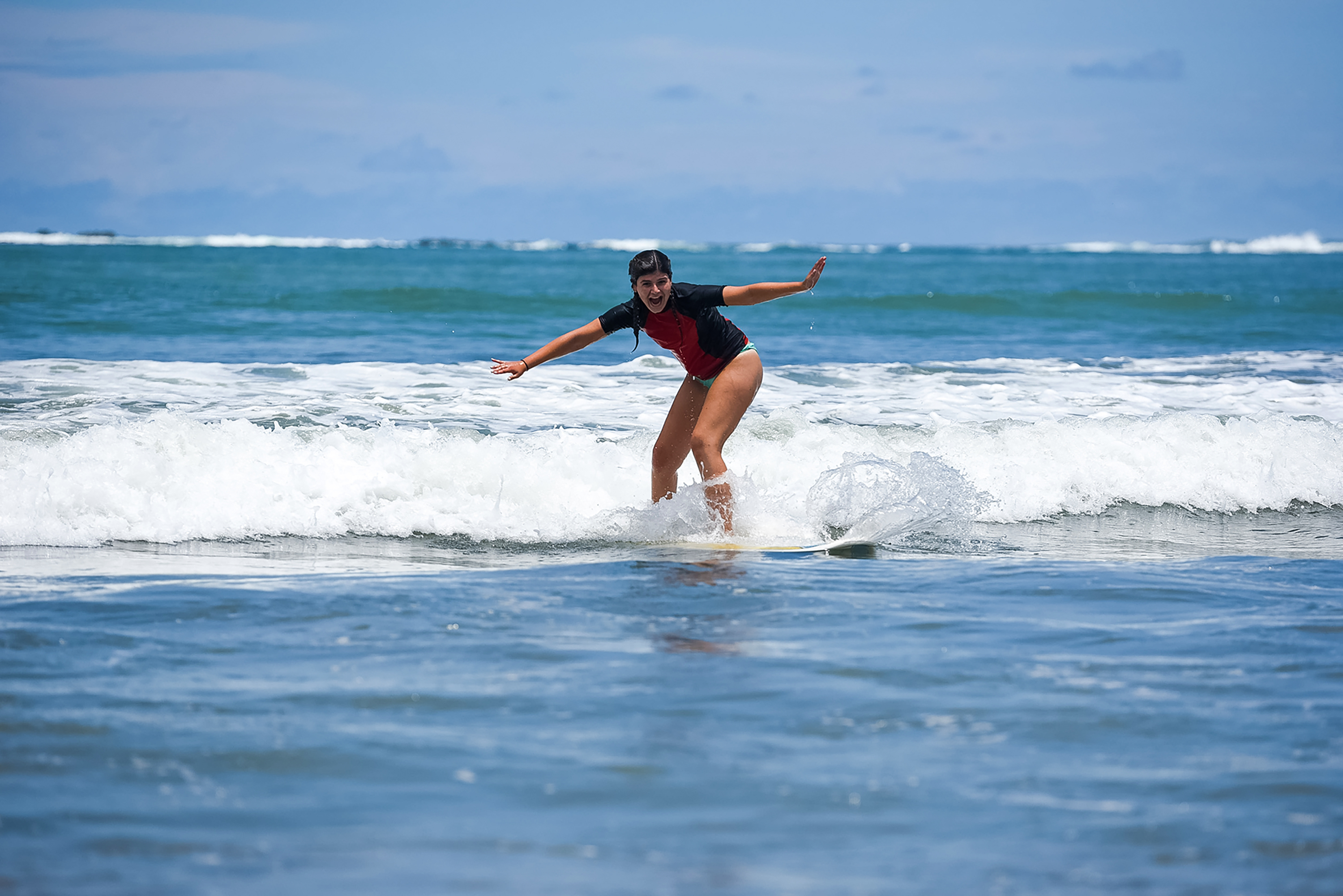 Teen travelers balance on a surfboard while riding a small wave