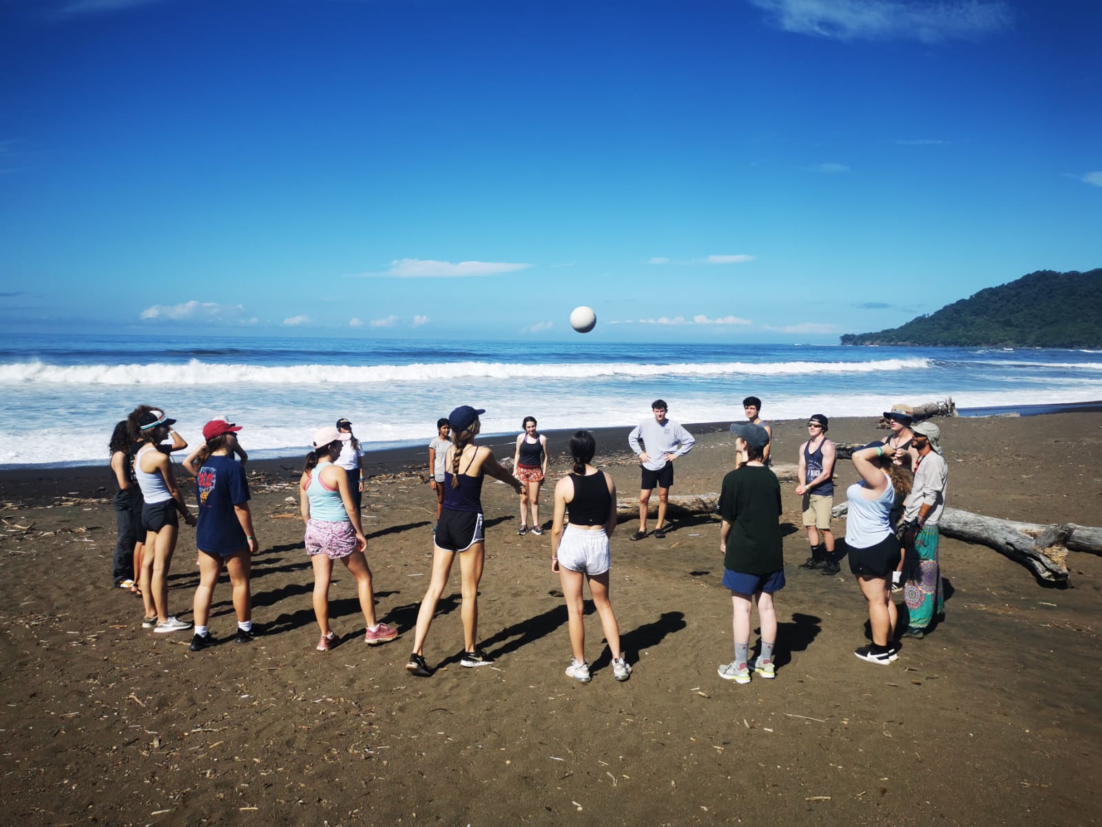 Students play a circle game with a ball on a dark-sand beach
