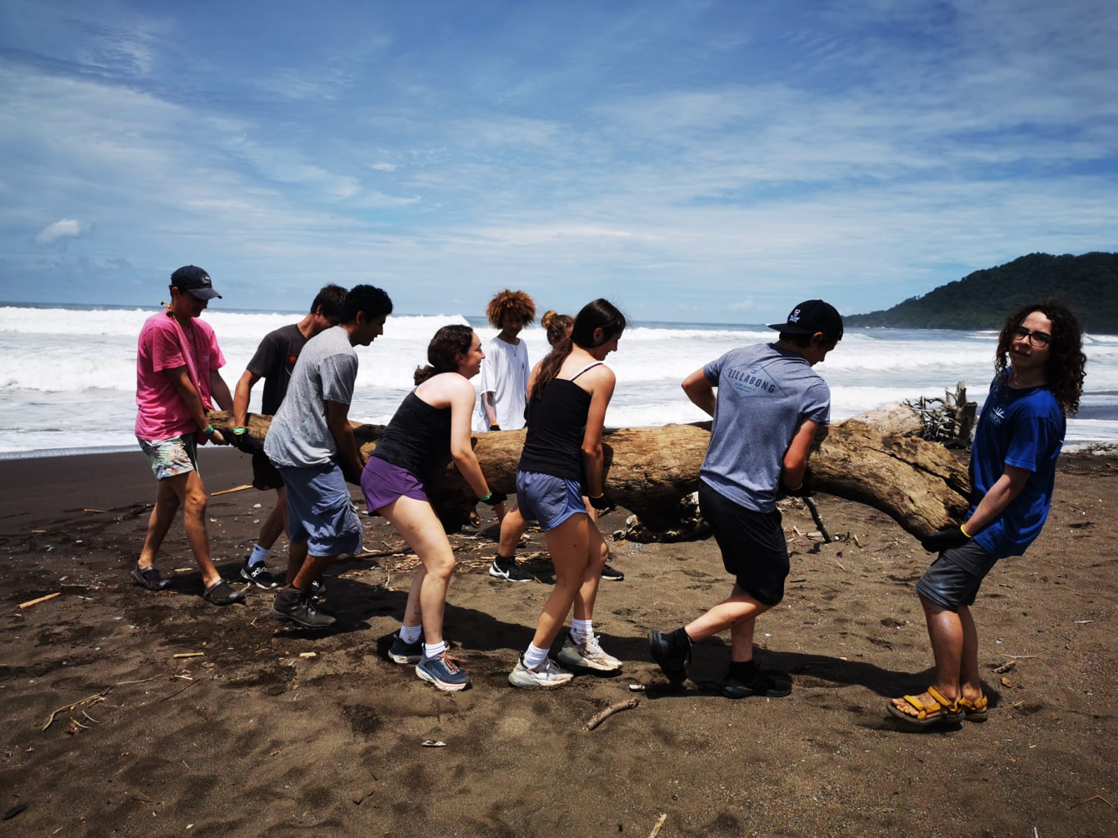 Students drag a large driftwood log across a black-sand beach