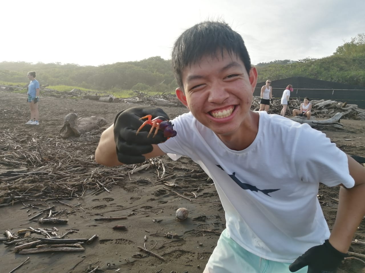 A student grins while holding a small crab on a driftwood-strewn beach