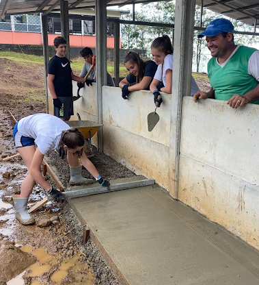On a school trip, a student smooths wet concrete while others watch from a wall on a service project