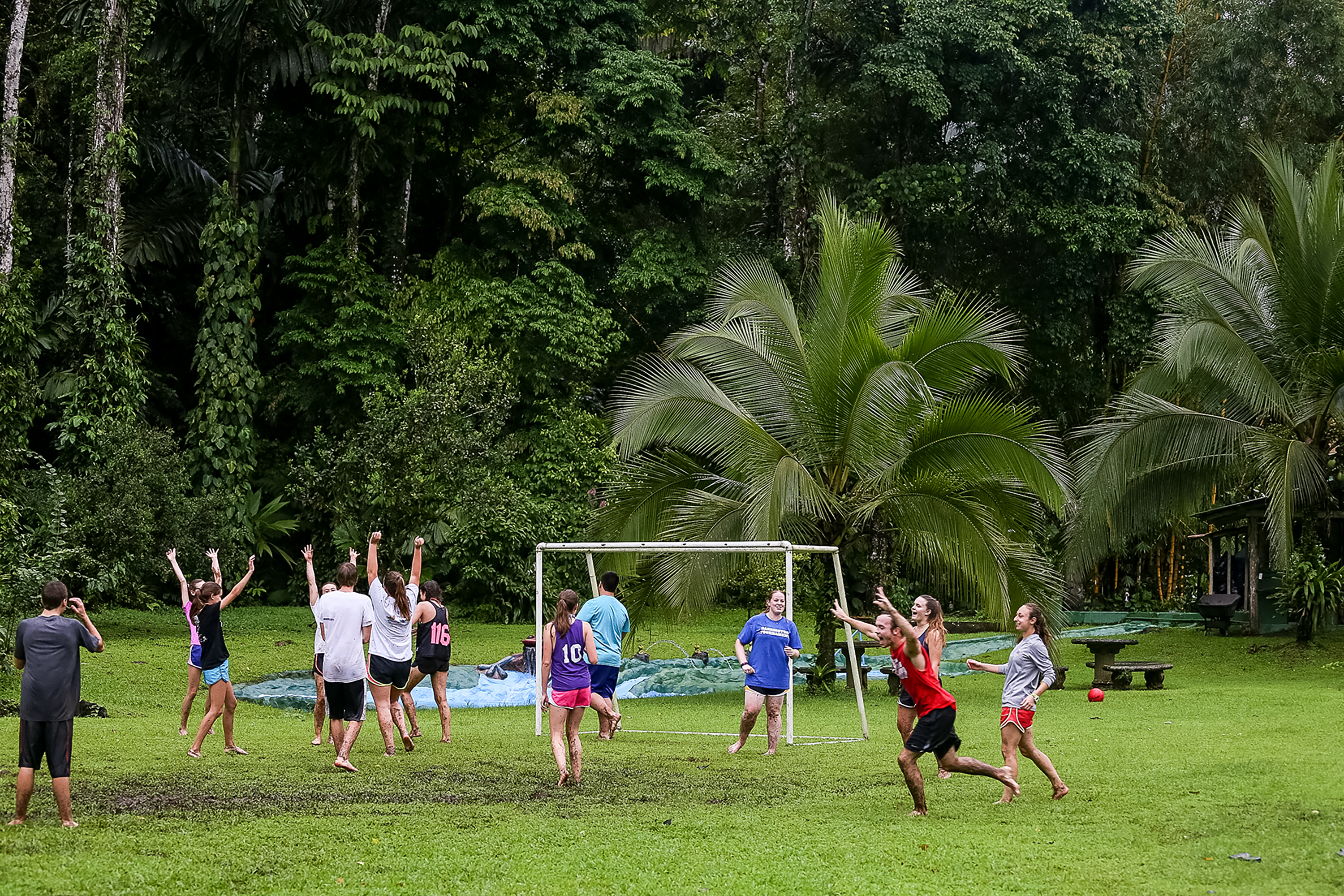 Students celebrate on a grassy field with a soccer goal amid palm trees and rainforest