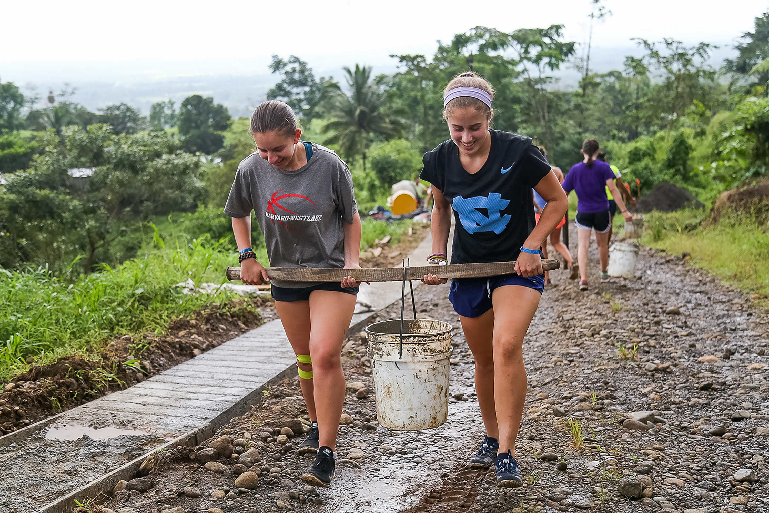 Students carry a bucket suspended from a pole along a muddy path on a community infrastructure project