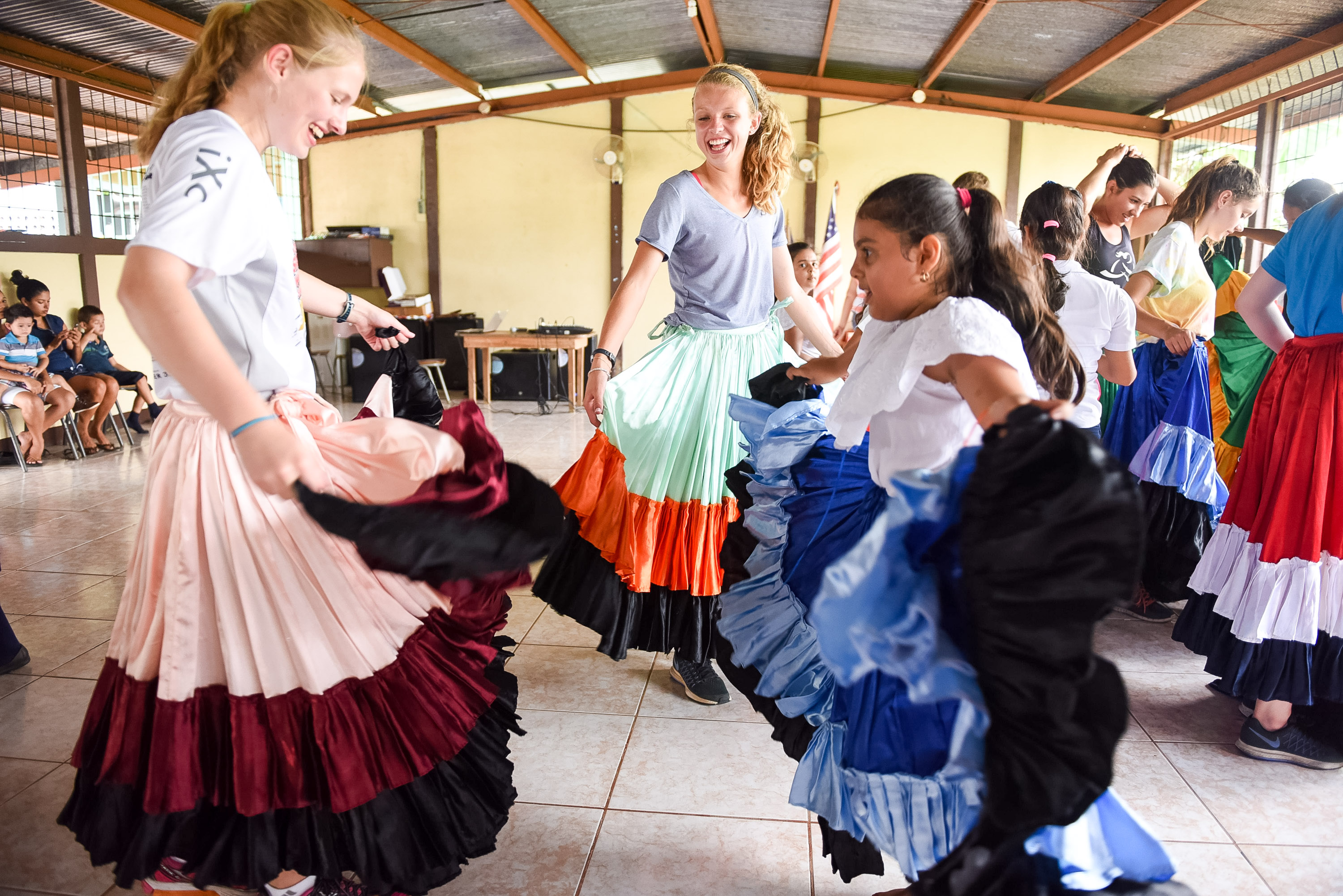 Students twirl colorful traditional skirts while dancing with a local girl indoors