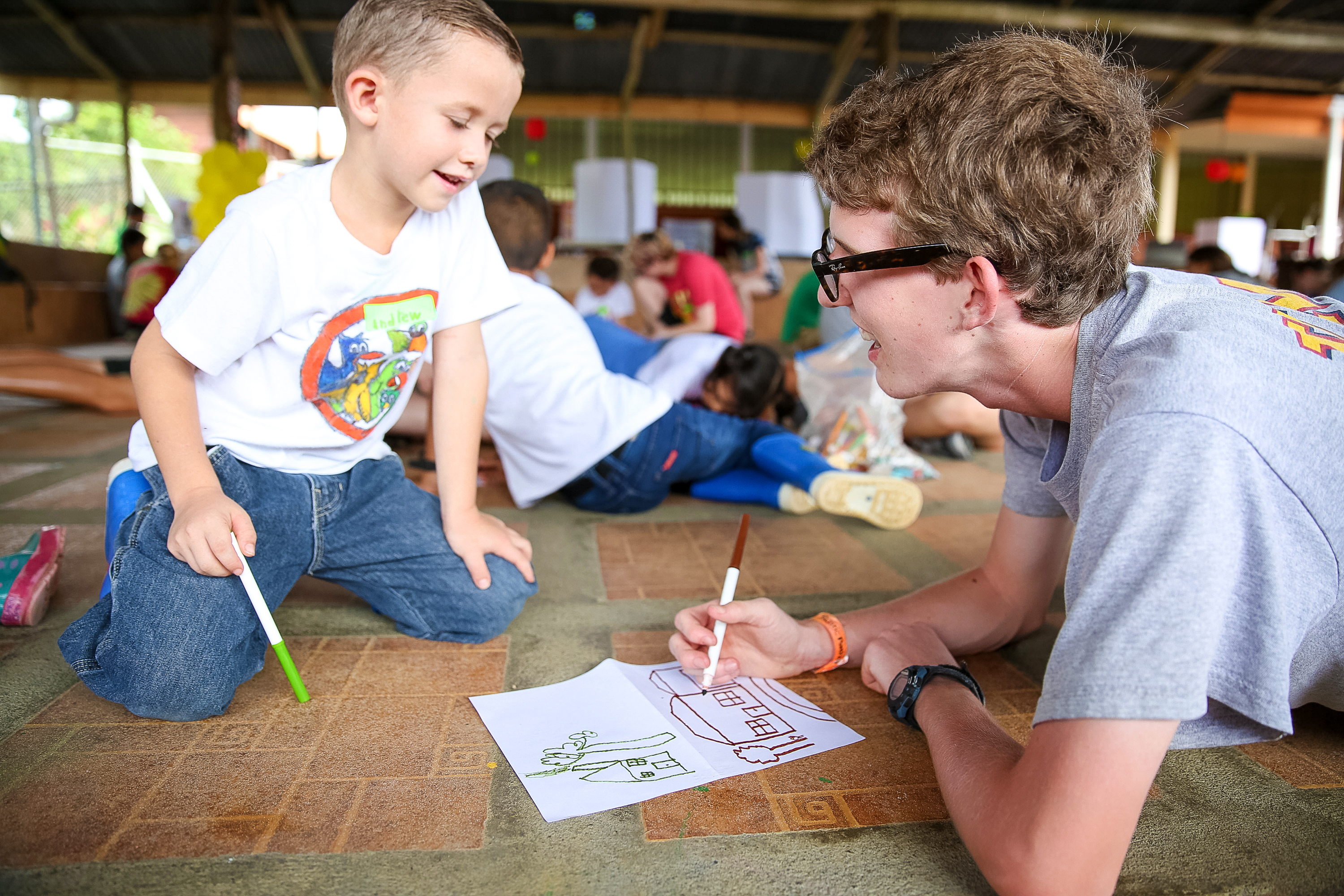 A student helps a young child color a drawing at an outdoor pavilion