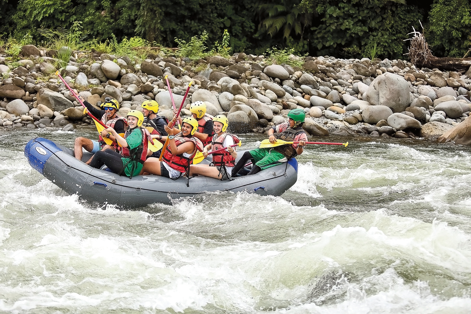 Students in helmets and life jackets paddle a raft through whitewater rapids lined with rocky banks