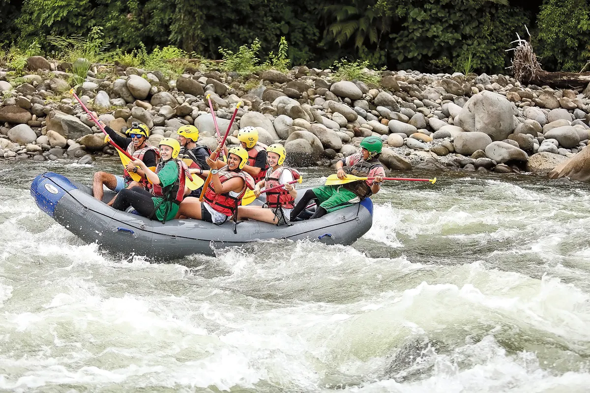 A school trip to Costa Rica rafts the Pacuare River, paddles raised as the raft drops through white water below a boulder bank