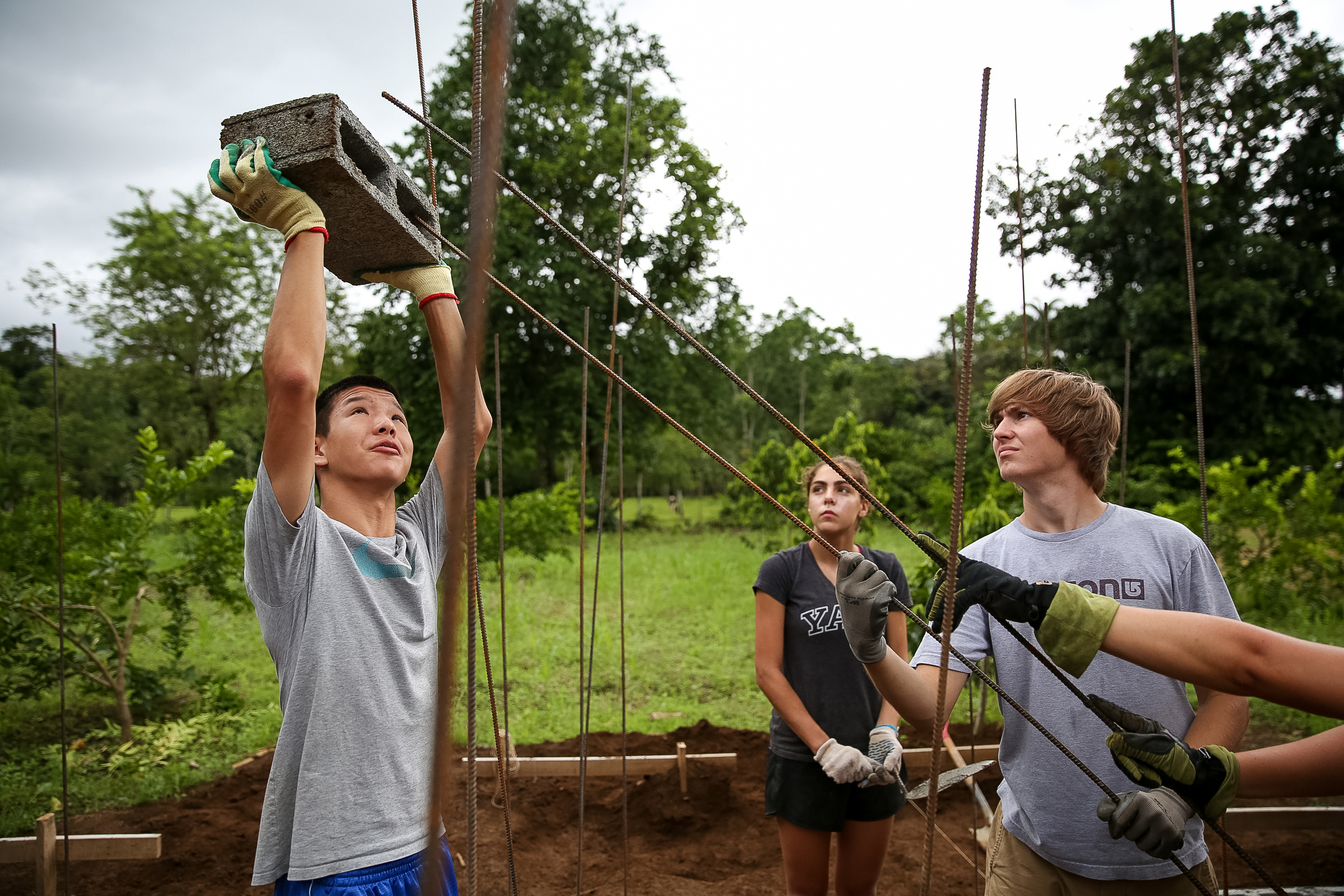 A student lifts a concrete block overhead while others hold rebar at a construction site