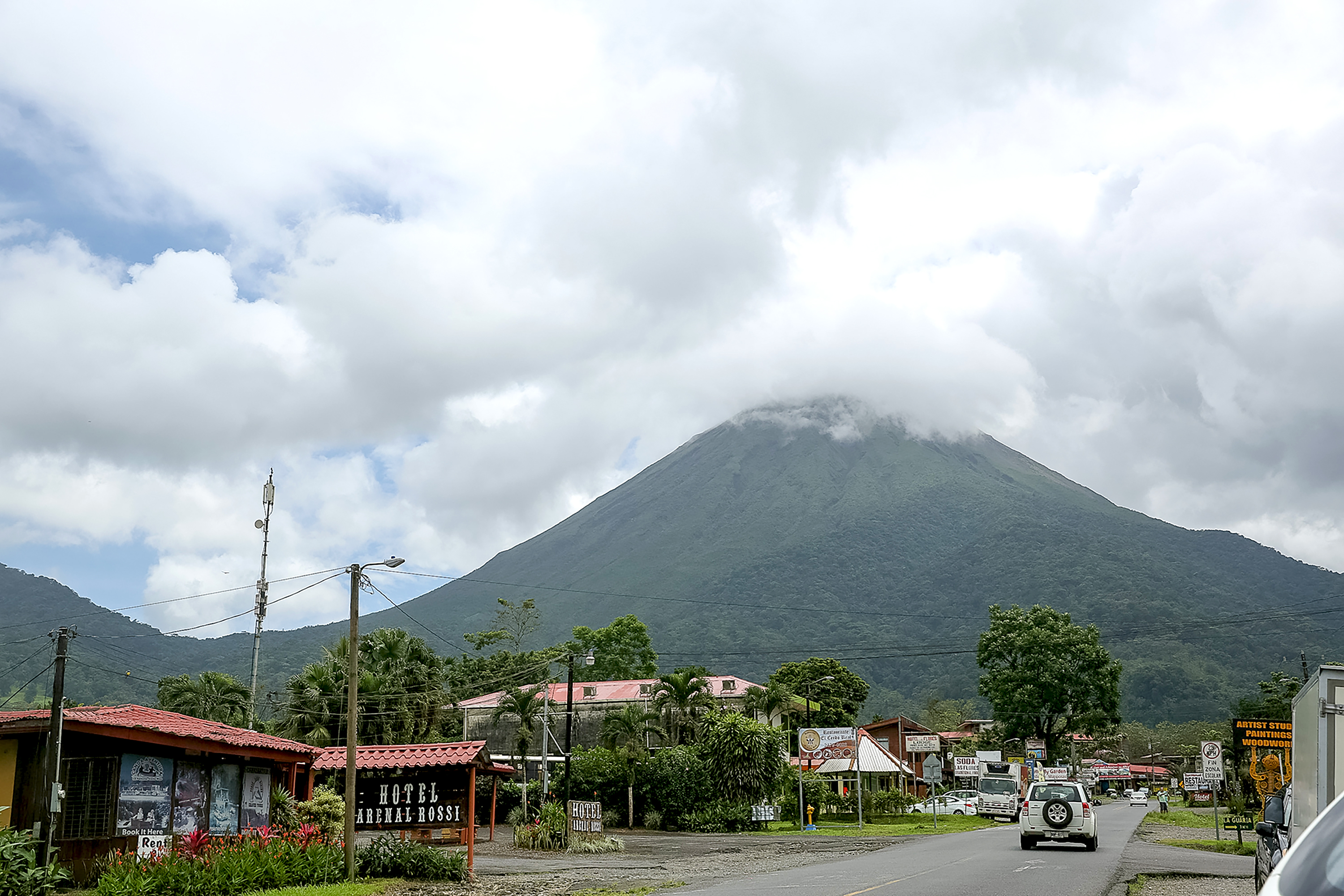 Teen travelers walk a small Costa Rican town street lined with shops beneath the rising Arenal Volcano