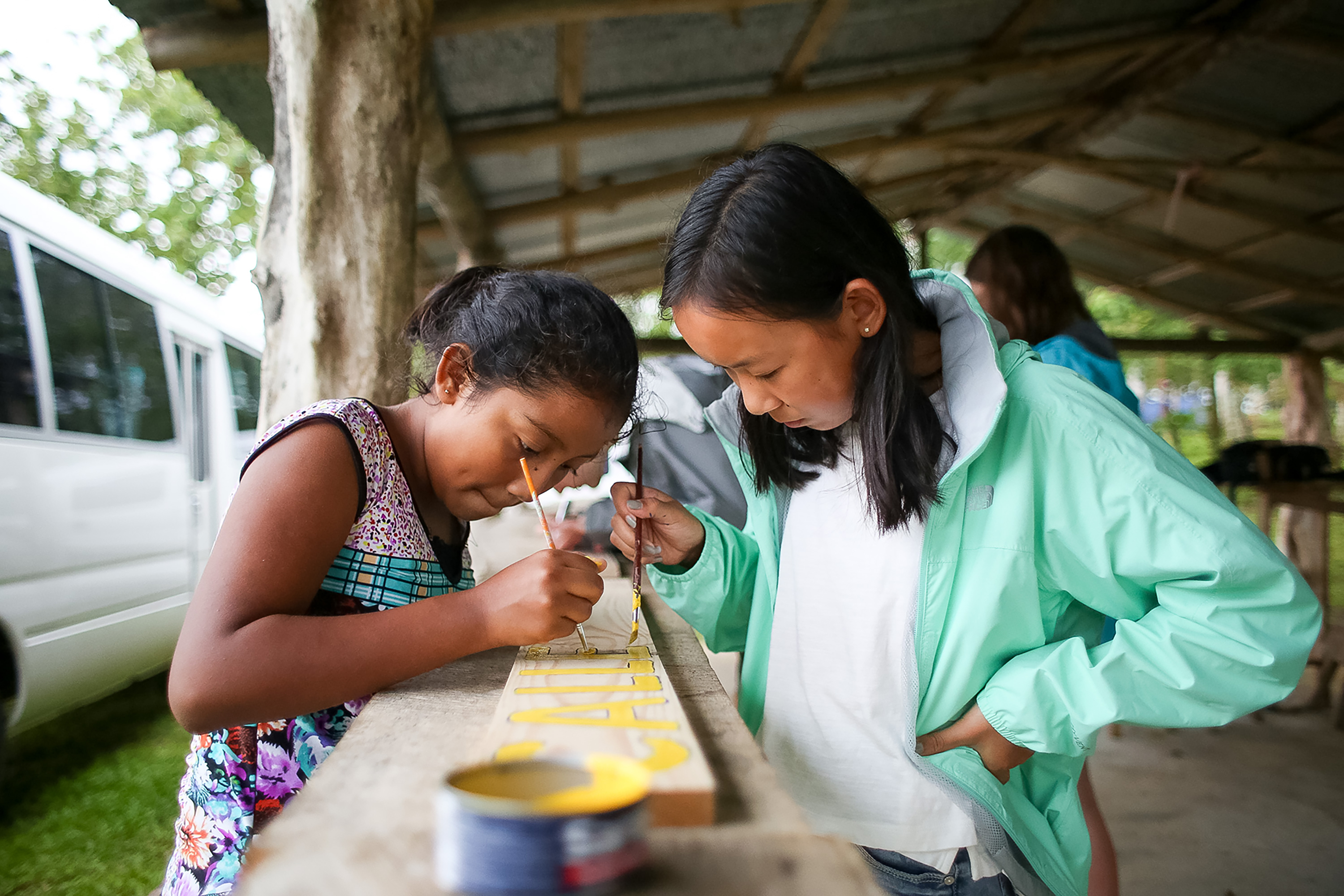 Students paint a wooden rail under an open-air shelter