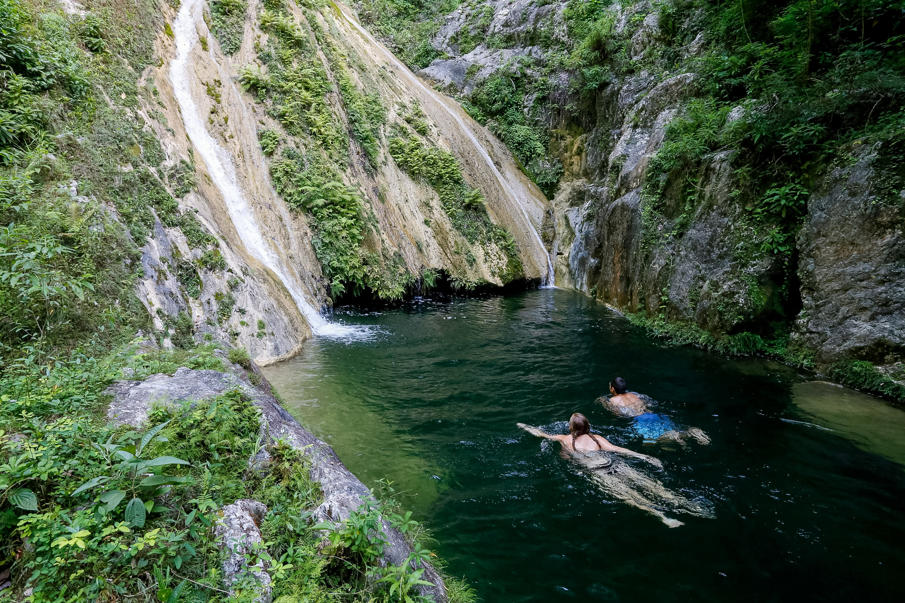 Students swim in a pool beneath a waterfall surrounded by cliffs and greenery