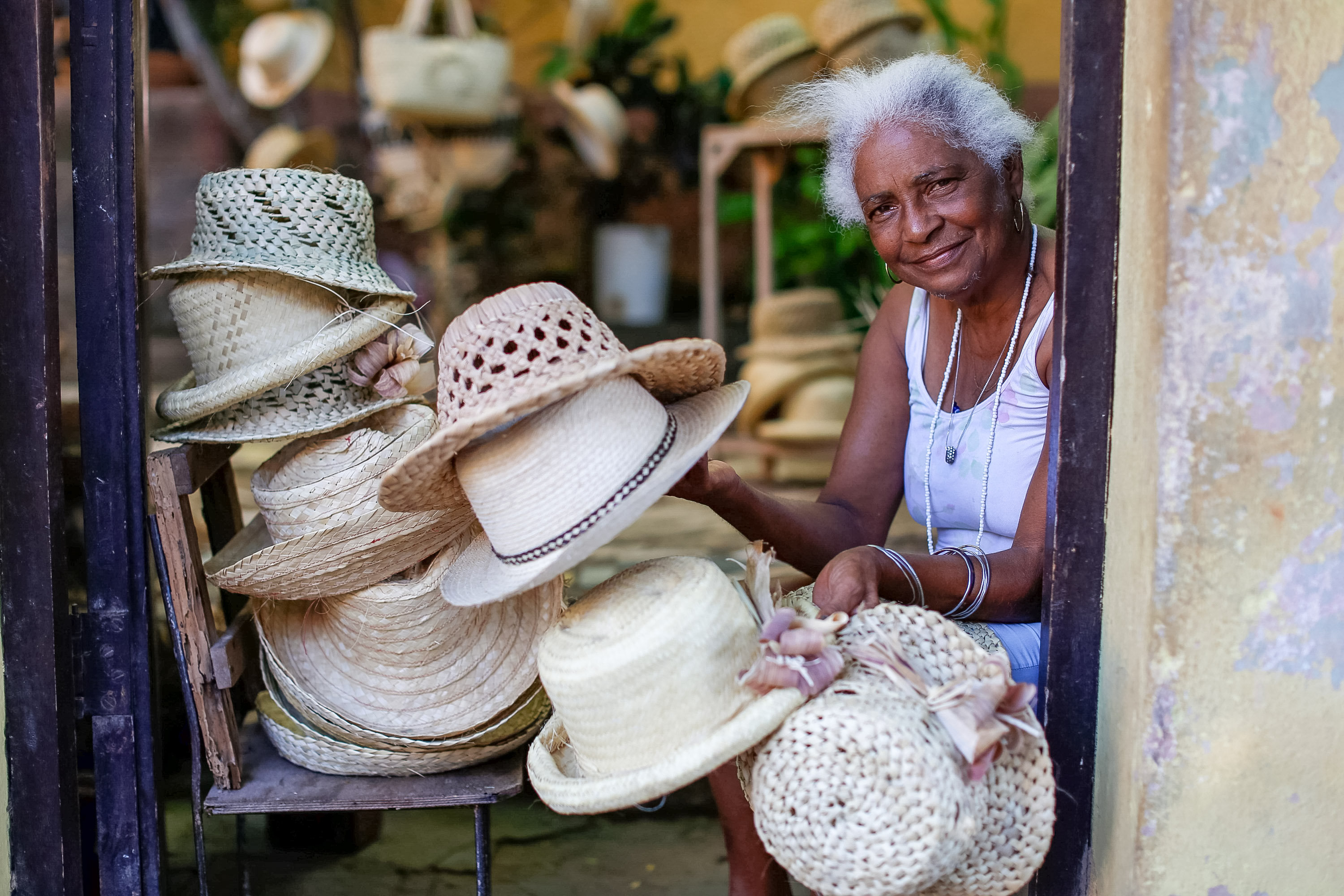 A smiling Cuban woman sits in a doorway beside stacks of handwoven straw hats.
