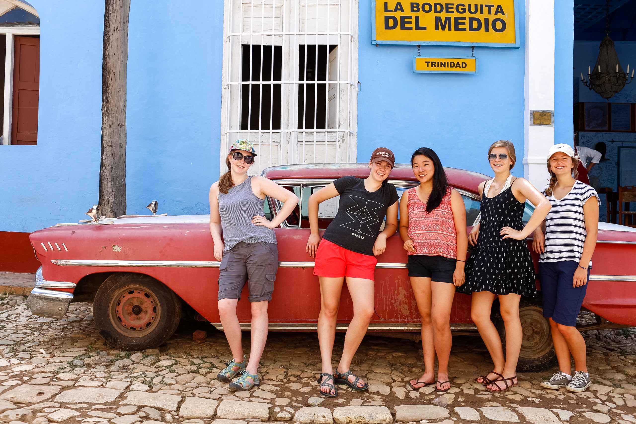 Four women posing in front of a red vintage car in front of colorful Cuban buildings.