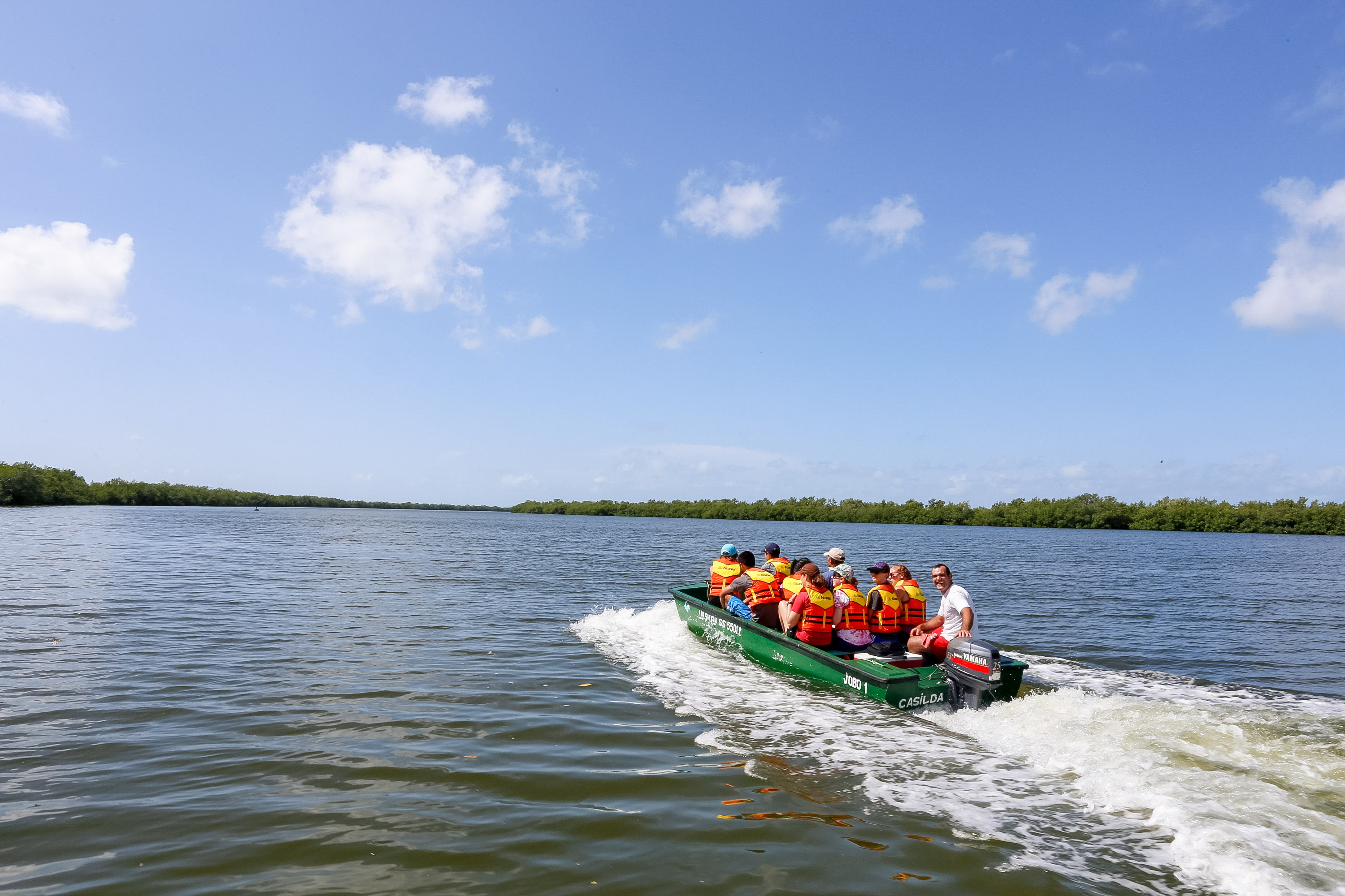 Students in life jackets ride a green boat across the water near mangroves