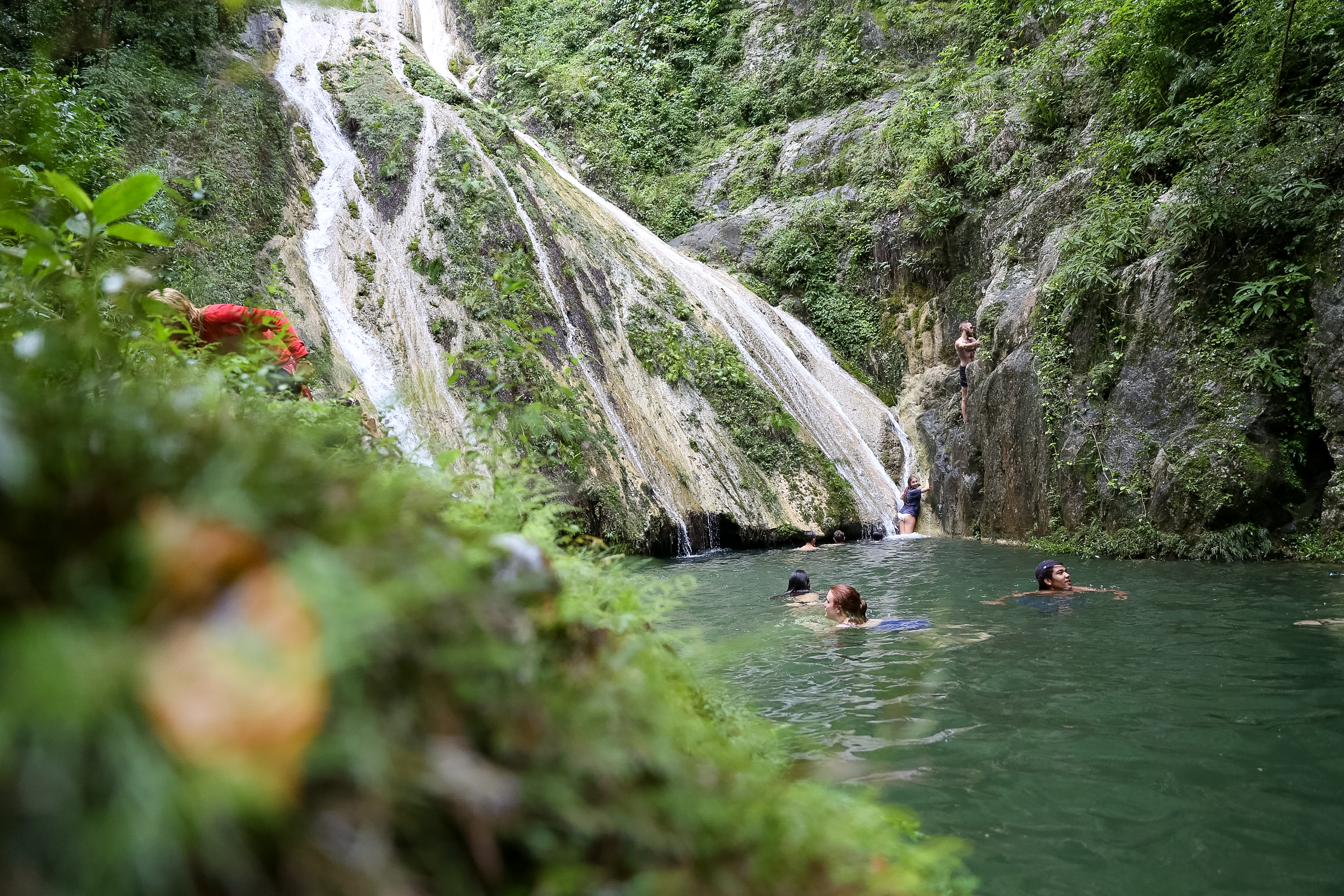 Students swim in a pool beneath a tall waterfall surrounded by lush cliffs