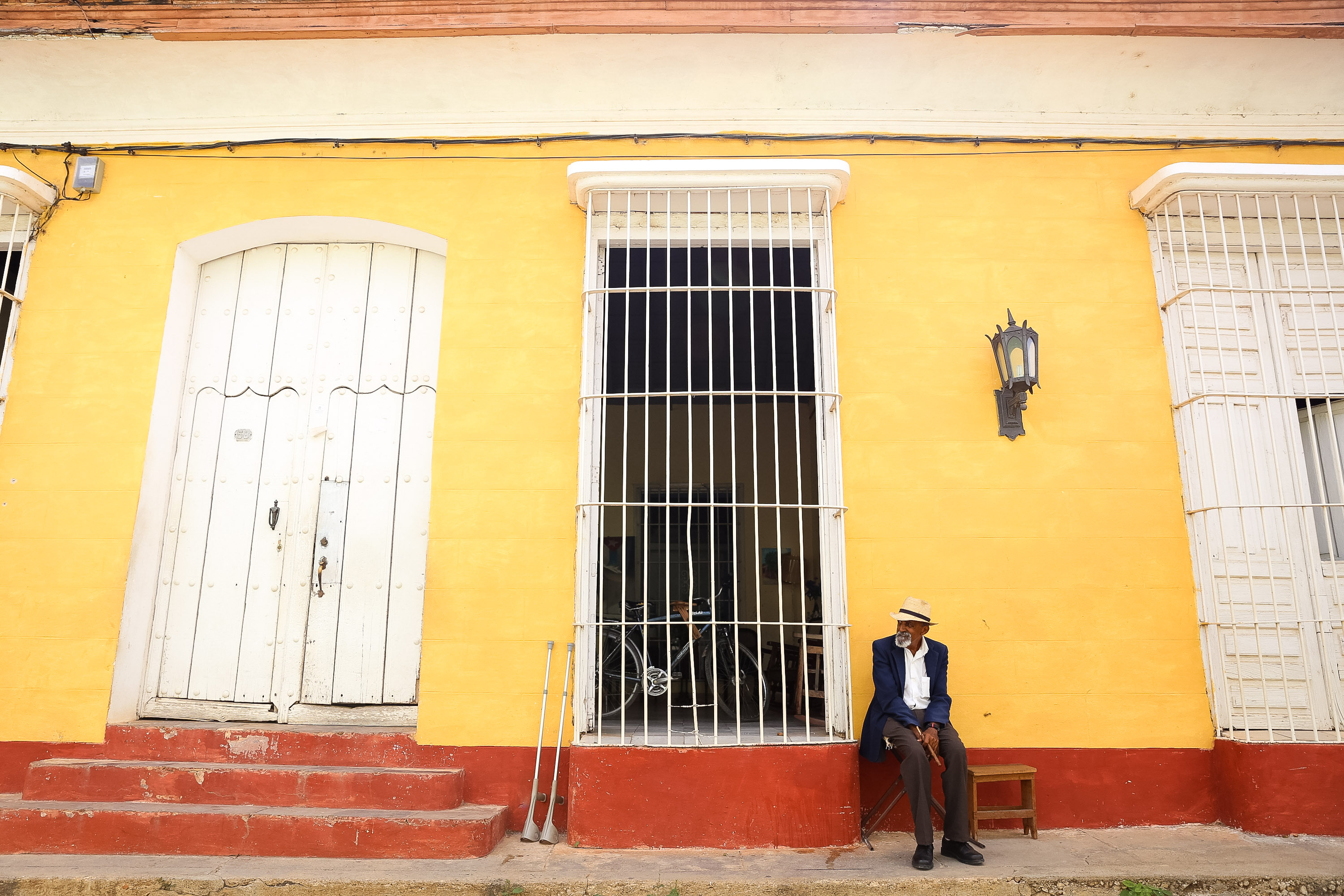 A man in a straw hat sits on a bench against a yellow colonial building