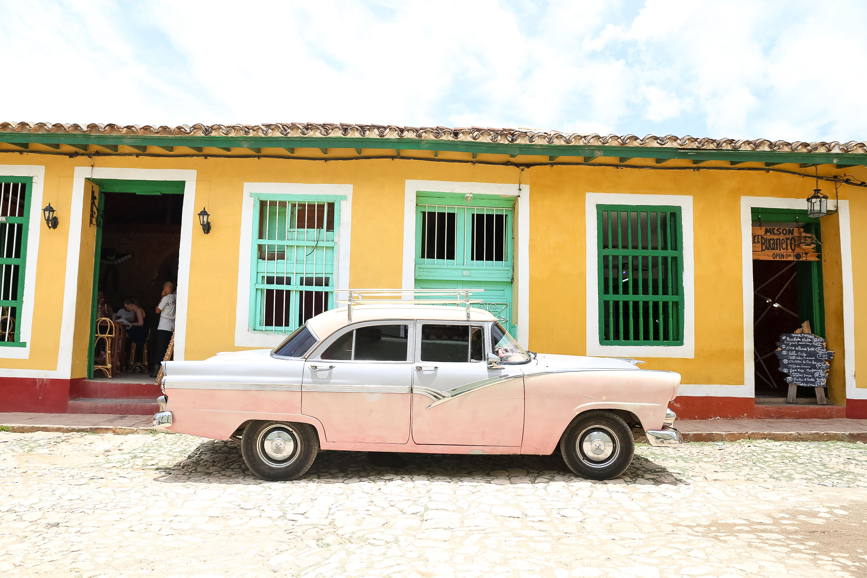 A pink 1950s American car parked outside a yellow colonial building on a cobblestone Trinidad street.