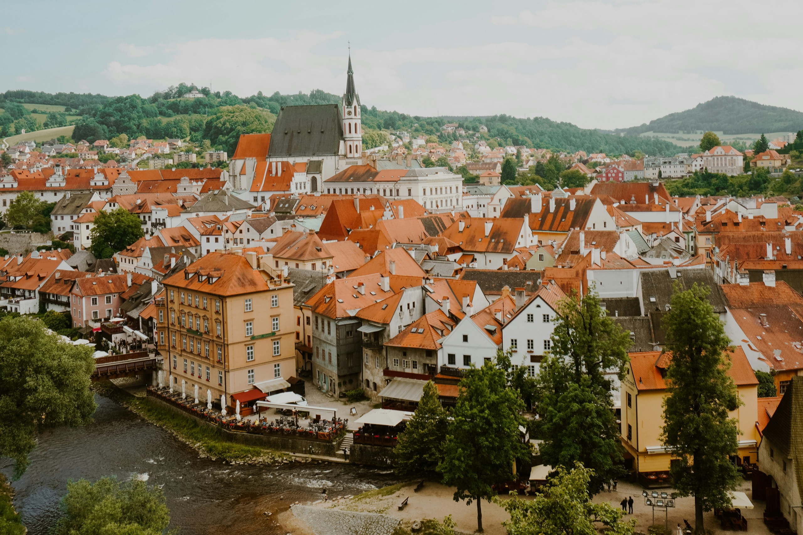 Red rooftops and the church spire of Český Krumlov above a bend of the Vltava, wooded hills behind the town.