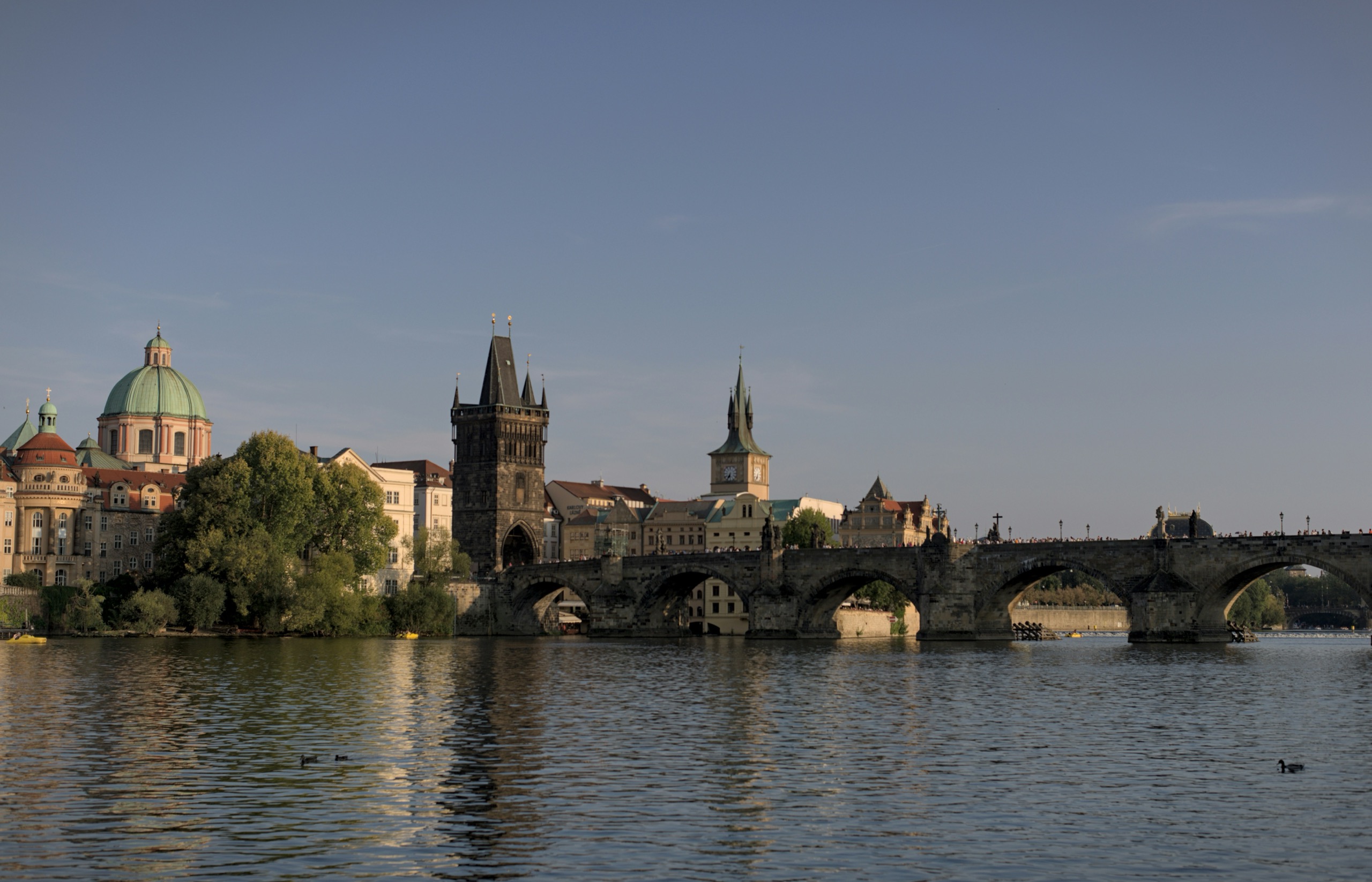 The stone arches of Charles Bridge seen from the river, the Old Town Bridge Tower and a green dome rising behind them.