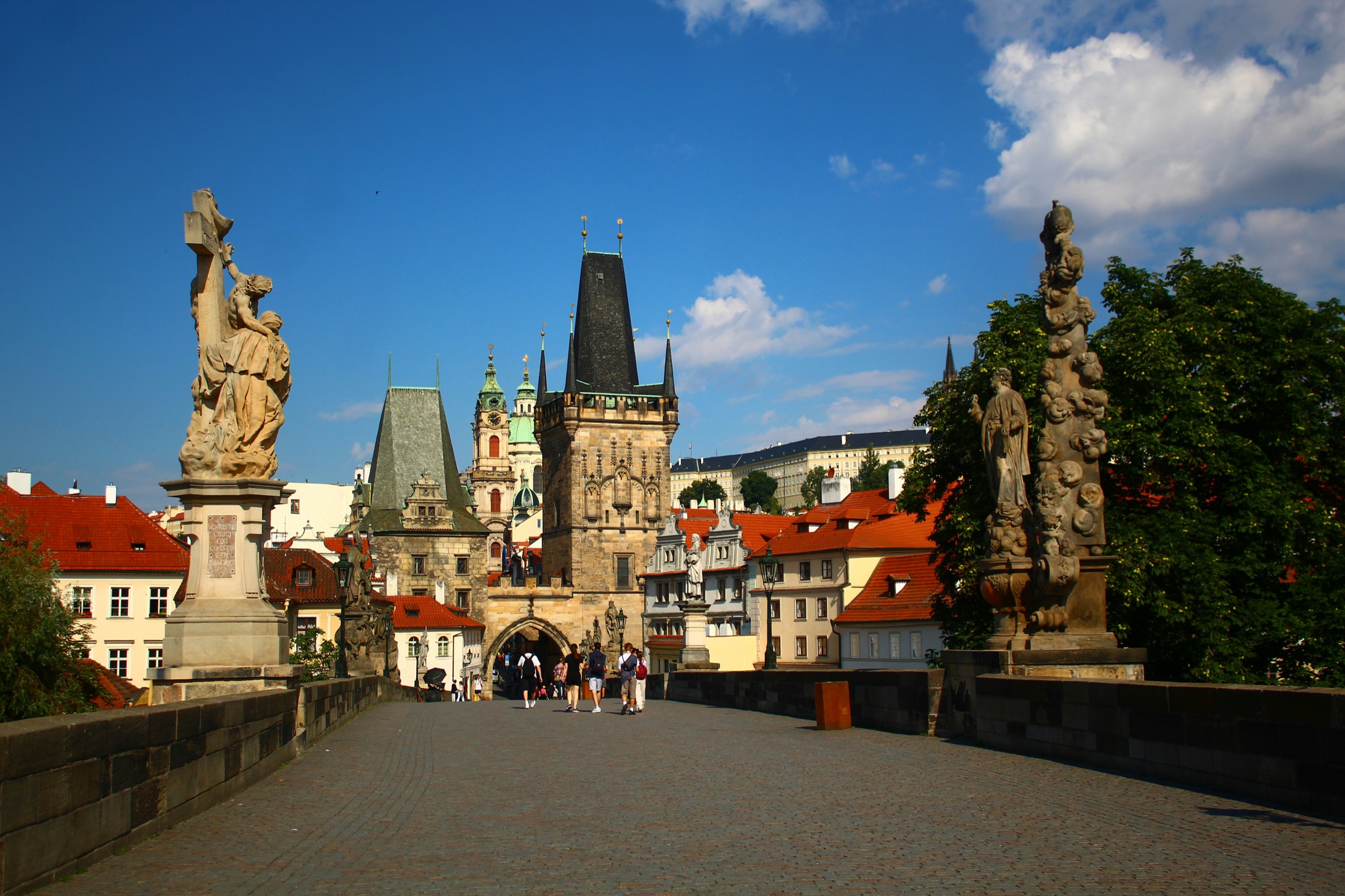 Baroque statues line Charles Bridge toward the Lesser Town bridge towers under a blue sky, a few walkers crossing the cobbles.