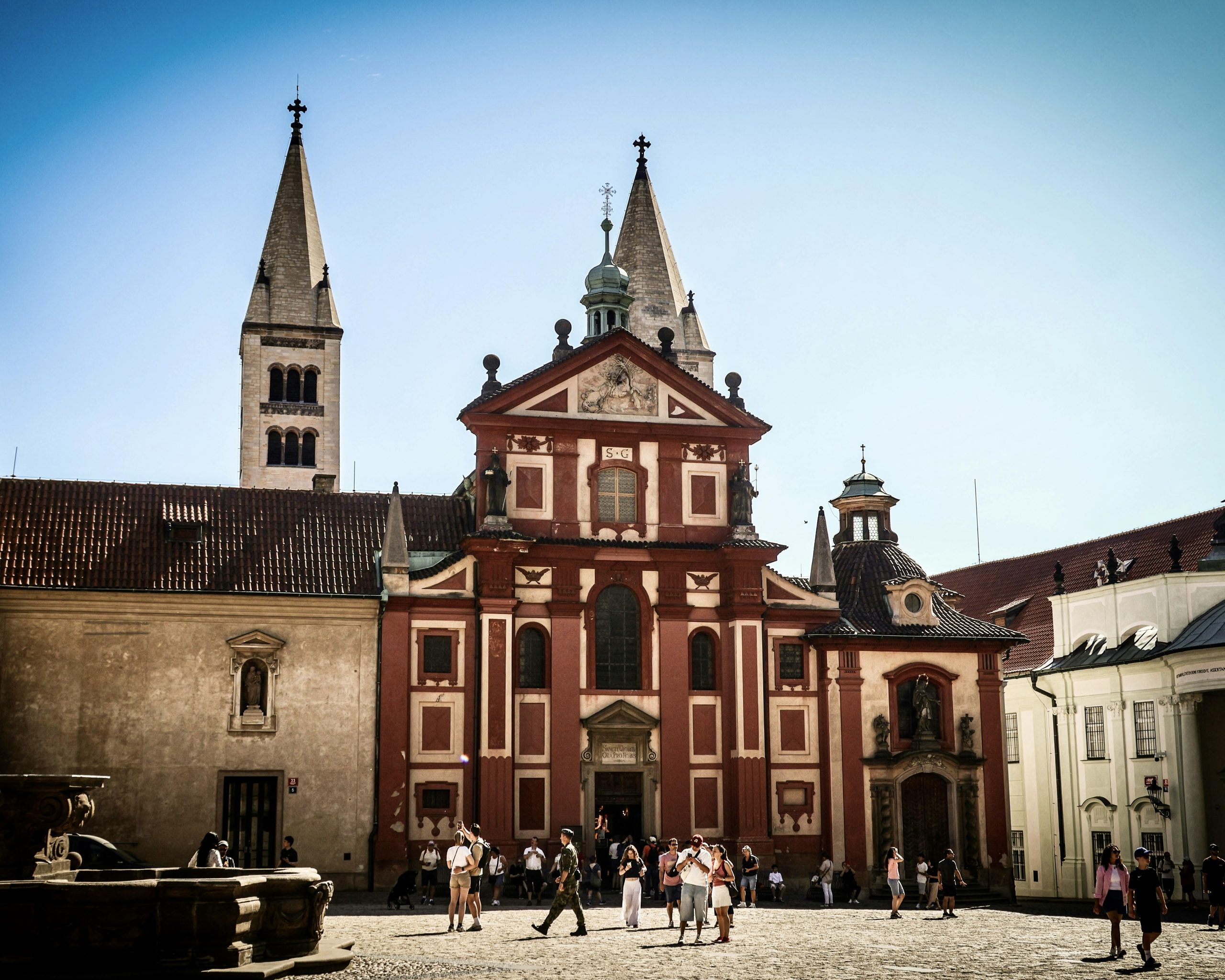 St. George's Basilica at Prague Castle, its red Baroque facade and twin Romanesque towers rising above a busy courtyard.