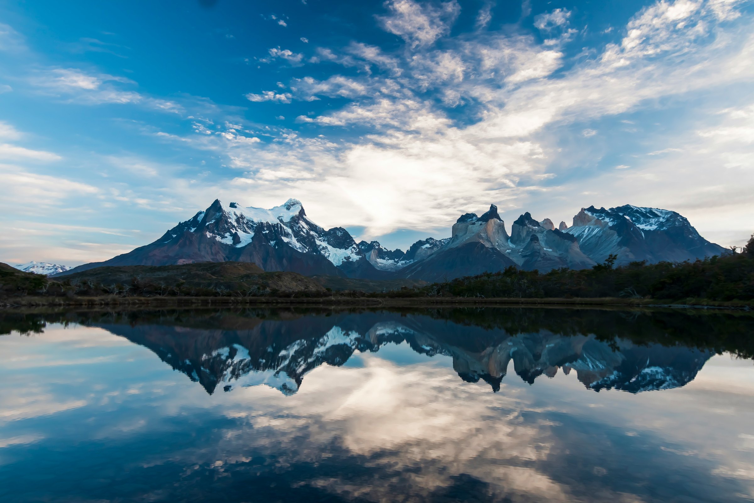Snow-streaked granite peaks under a bright, cloud-streaked sky, mirrored almost exactly in a still lake with a low line of scrub along the far shore.