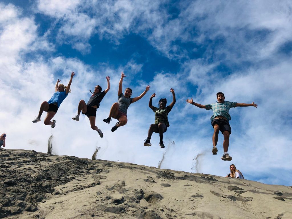 People jumping off a sand dune under a bright blue sky.