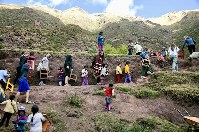 Teen travelers help local children carry small wooden chairs up a terraced hillside in a village in Peru