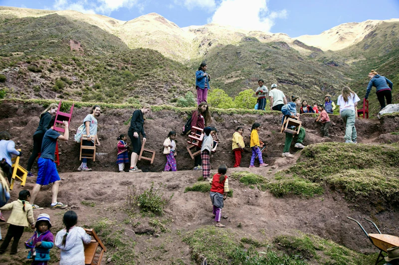 Teen travelers help local children carry small wooden chairs up a terraced hillside in a village in Peru