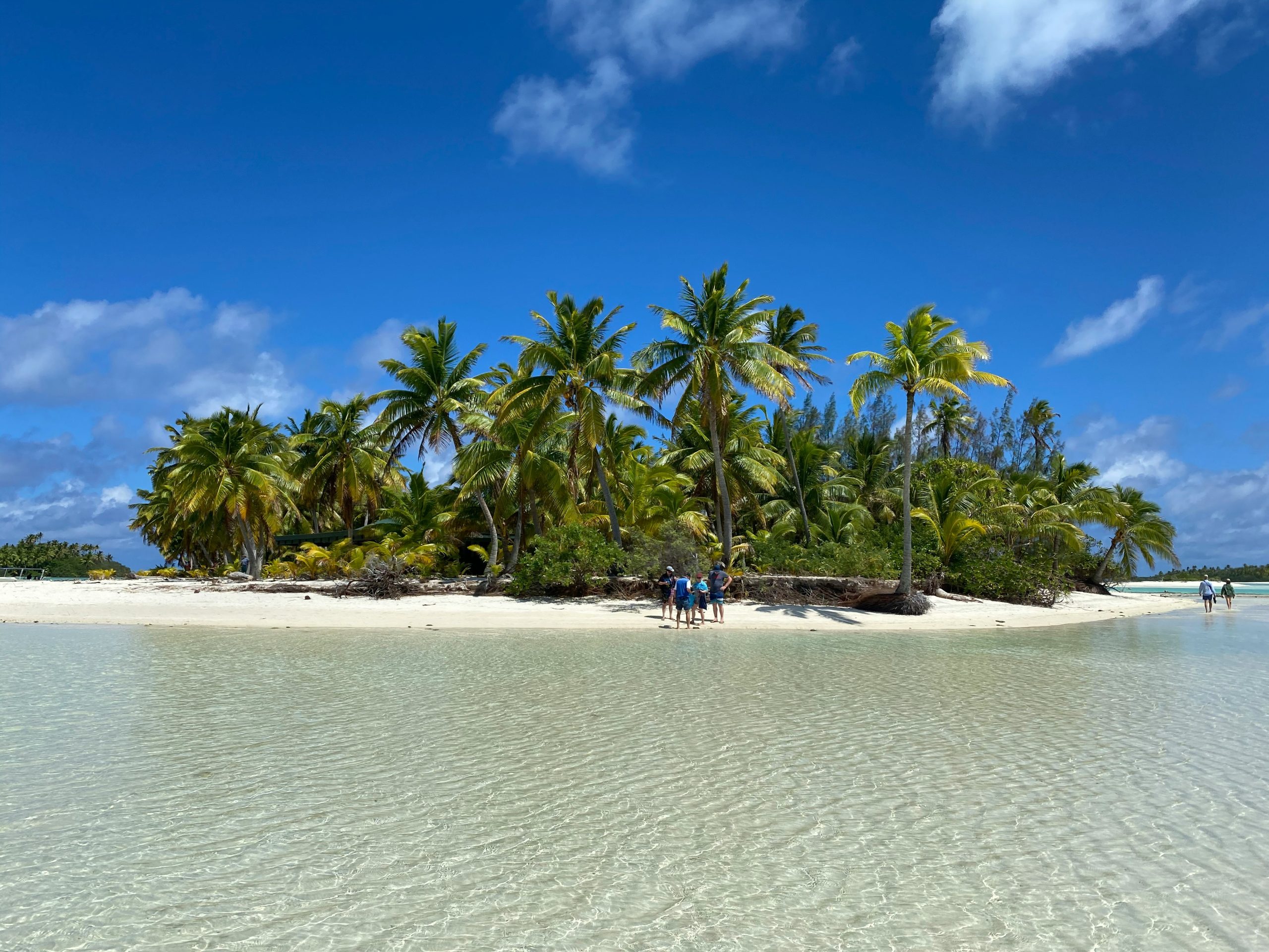 A small group stands on a white-sand beach beside a dense palm-covered motu, with clear turquoise lagoon water in the foreground.