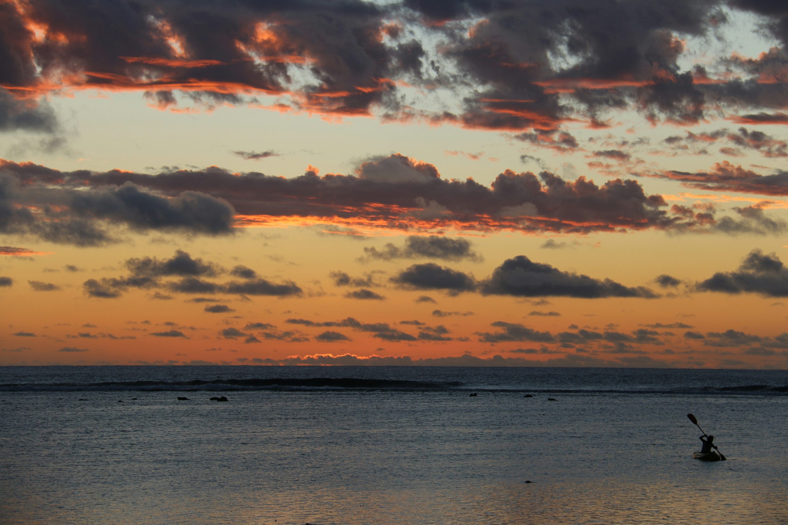 A person paddles a kayak across calm water at dusk, beneath a sky streaked with orange and pink clouds.