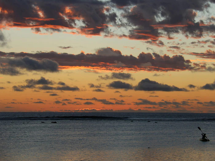 A person paddles a kayak across calm water at dusk, beneath a sky streaked with orange and pink clouds.