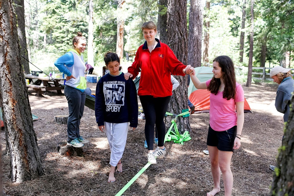 This photo shows summer campers helping each other balance on a slackline stretched between trees.