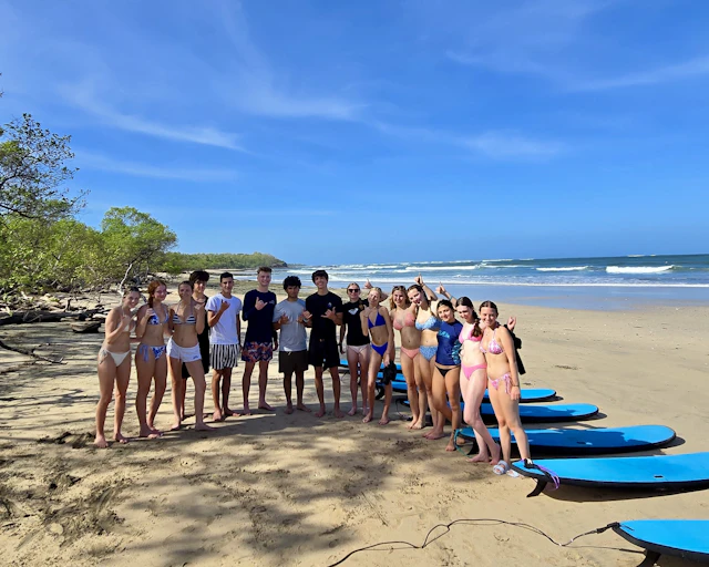 Students in swimsuits stand behind a row of blue surfboards on a beach in Costa Rica