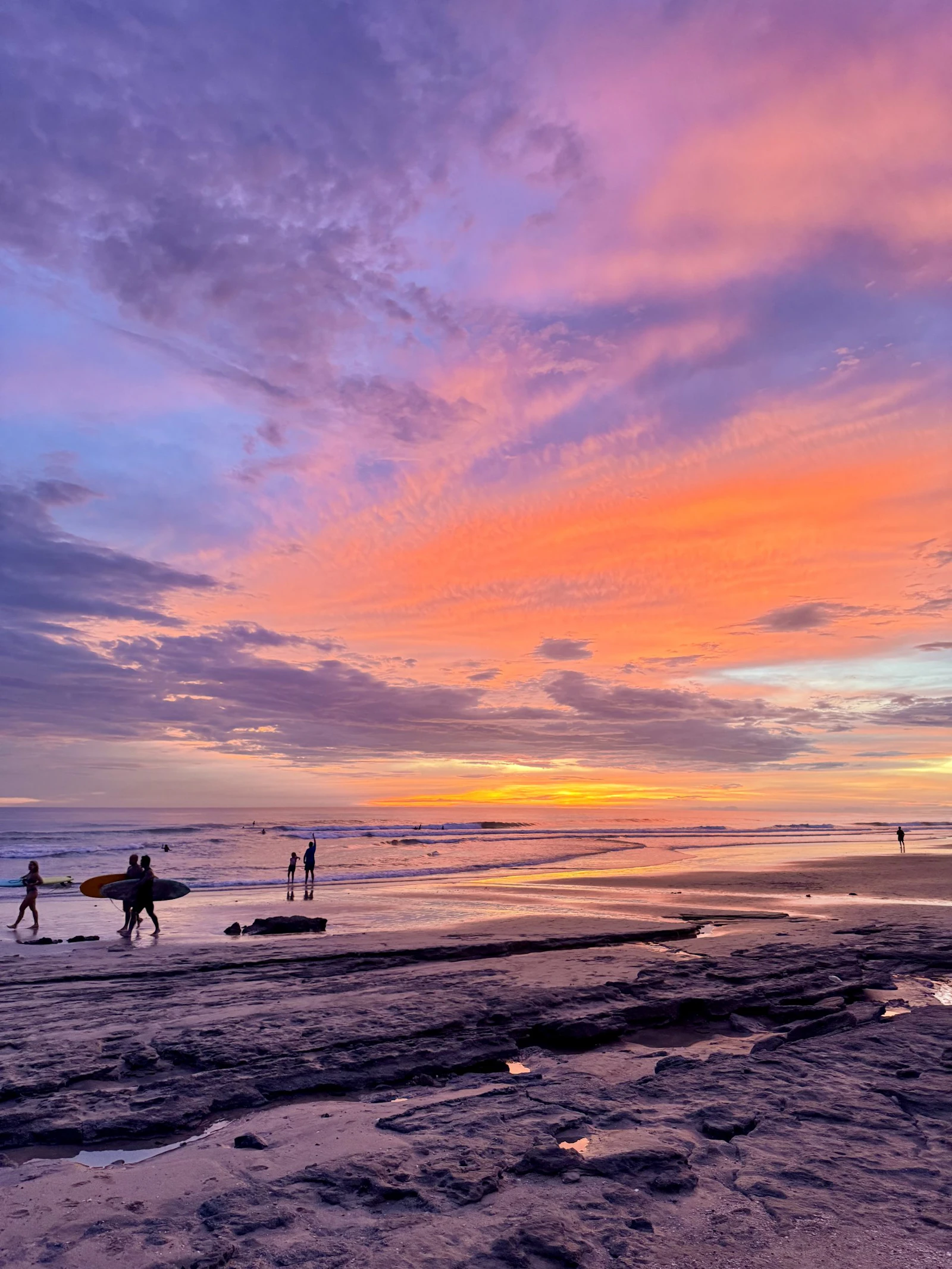 Students carry surfboards along the shore under an orange and pink sunset in Costa Rica
