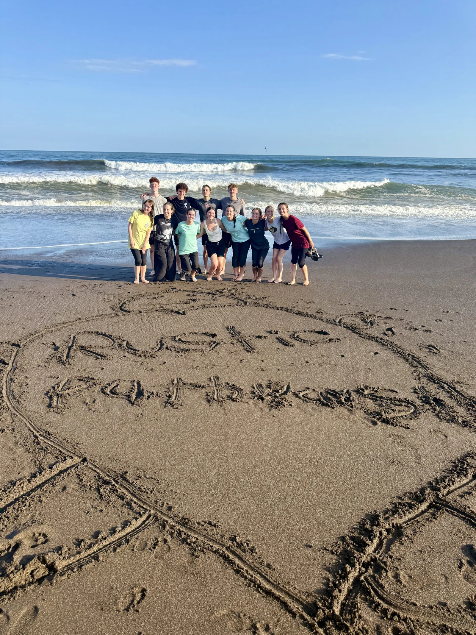 Students stand on a dark sand beach in Costa Rica behind the words Rustic Pathways written in the sand