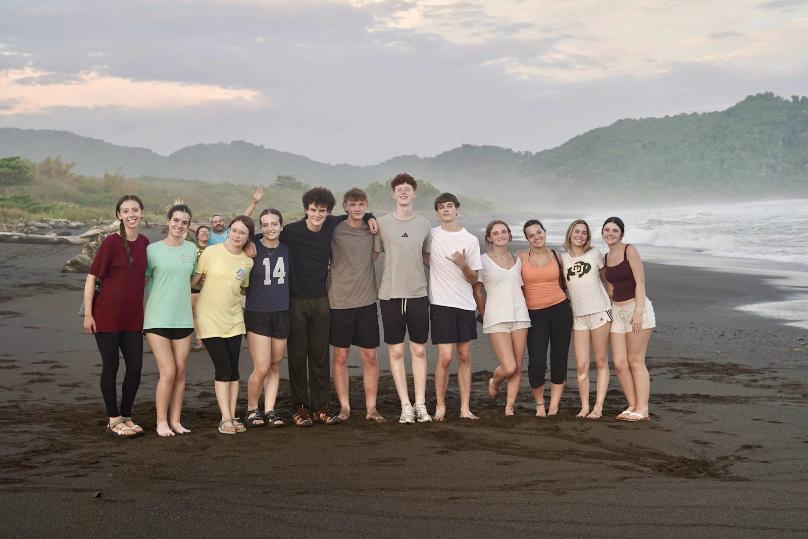 A Rustic Pathways group stands together on a dark sand beach below forested hills in Costa Rica