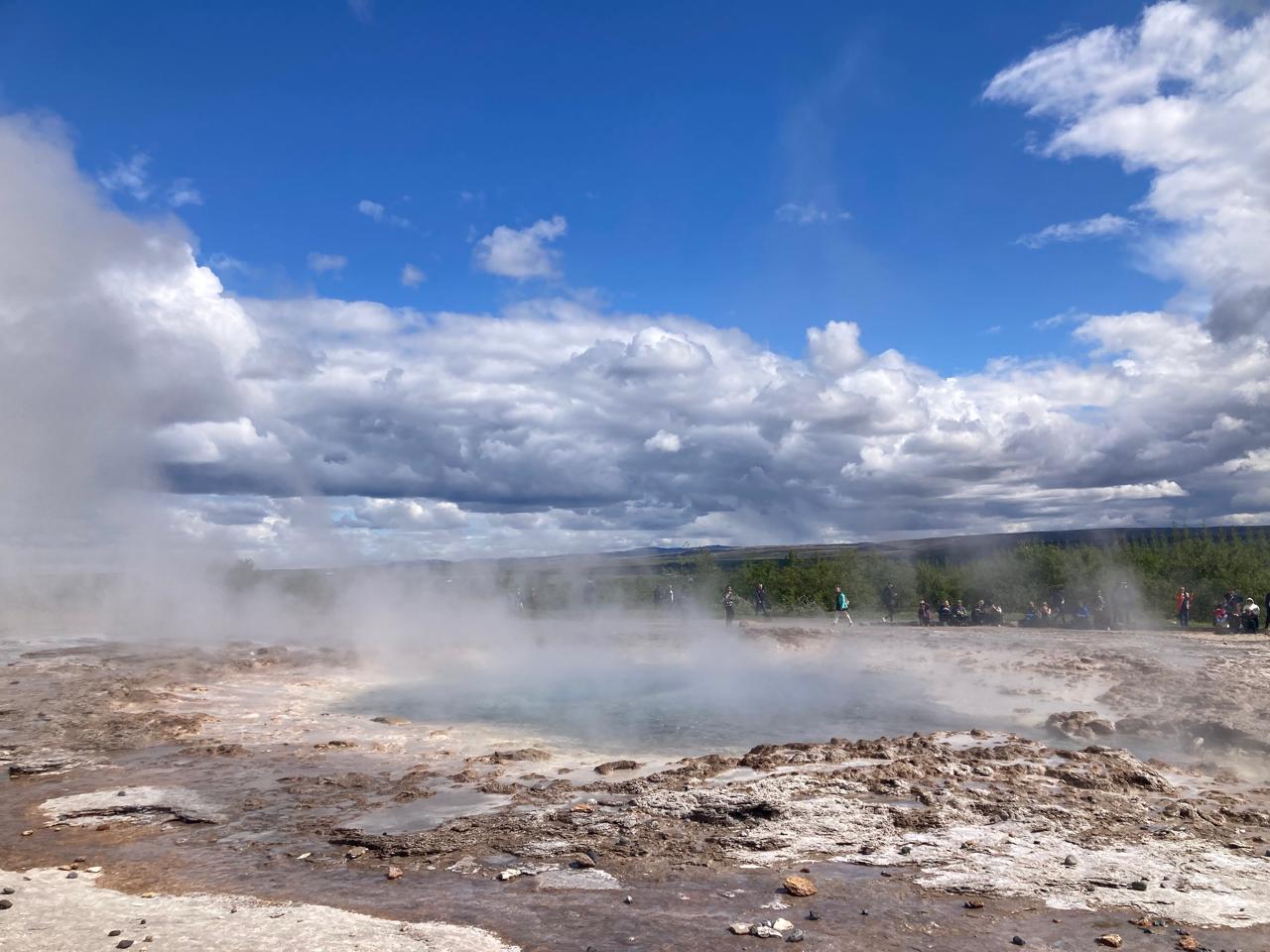 Steam rises from a geothermal geyser pool under a blue sky with scattered clouds