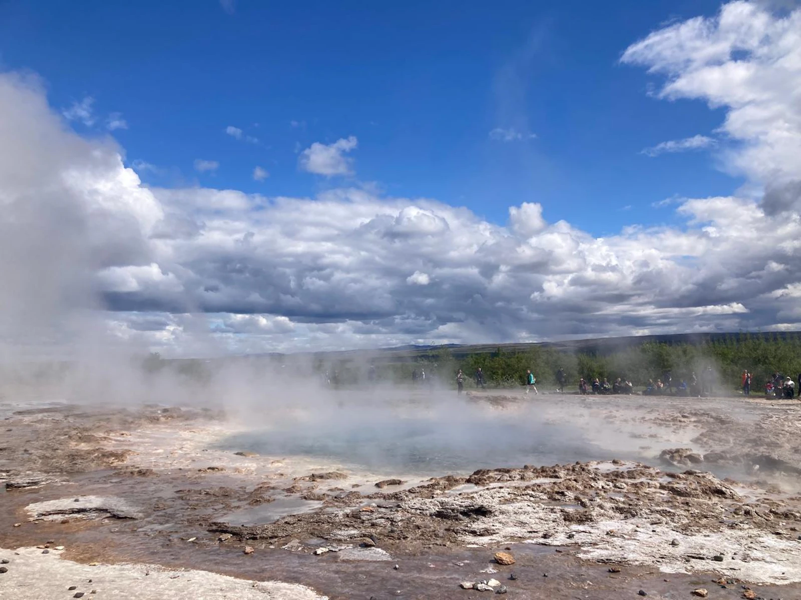 Steam drifts across a geothermal field of bubbling pools and pale mineral crust under a blue sky