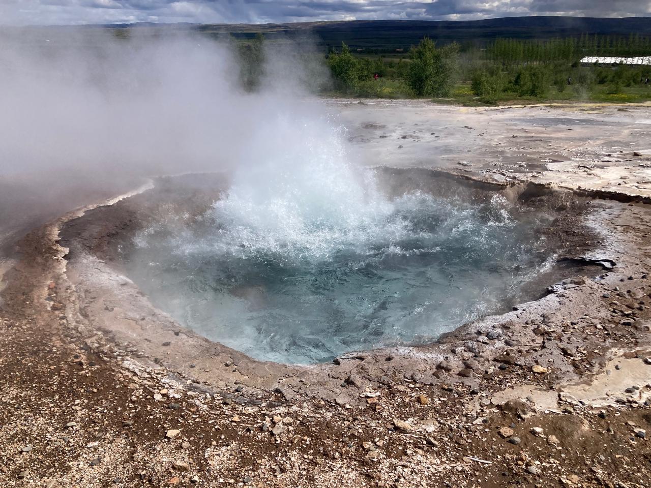 A geyser erupts with steam and water inside a mineral-rimmed pool