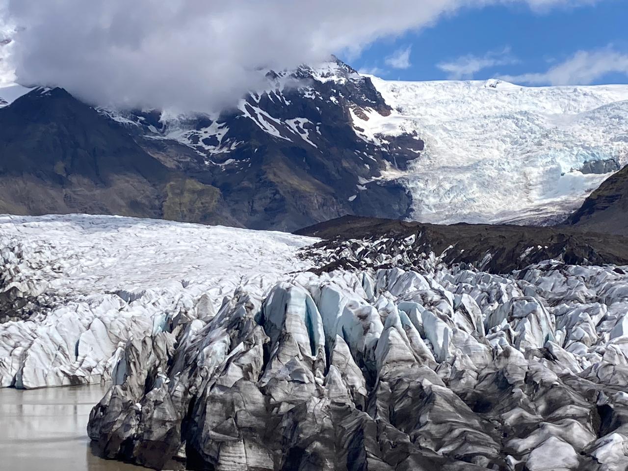 Jagged glacier ice formations rise near a lagoon with snow-capped mountains beyond