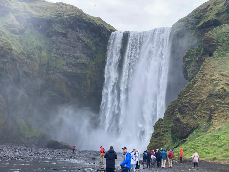 A group of travelers stands on black gravel at the foot of a tall waterfall pouring between steep green cliffs