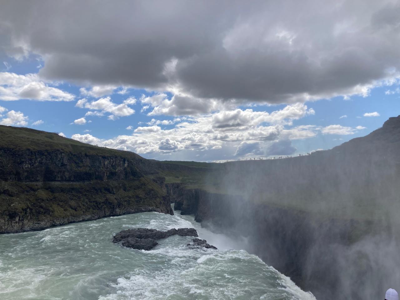 Mist rises over a powerful waterfall surrounded by rugged cliffs under a cloudy sky