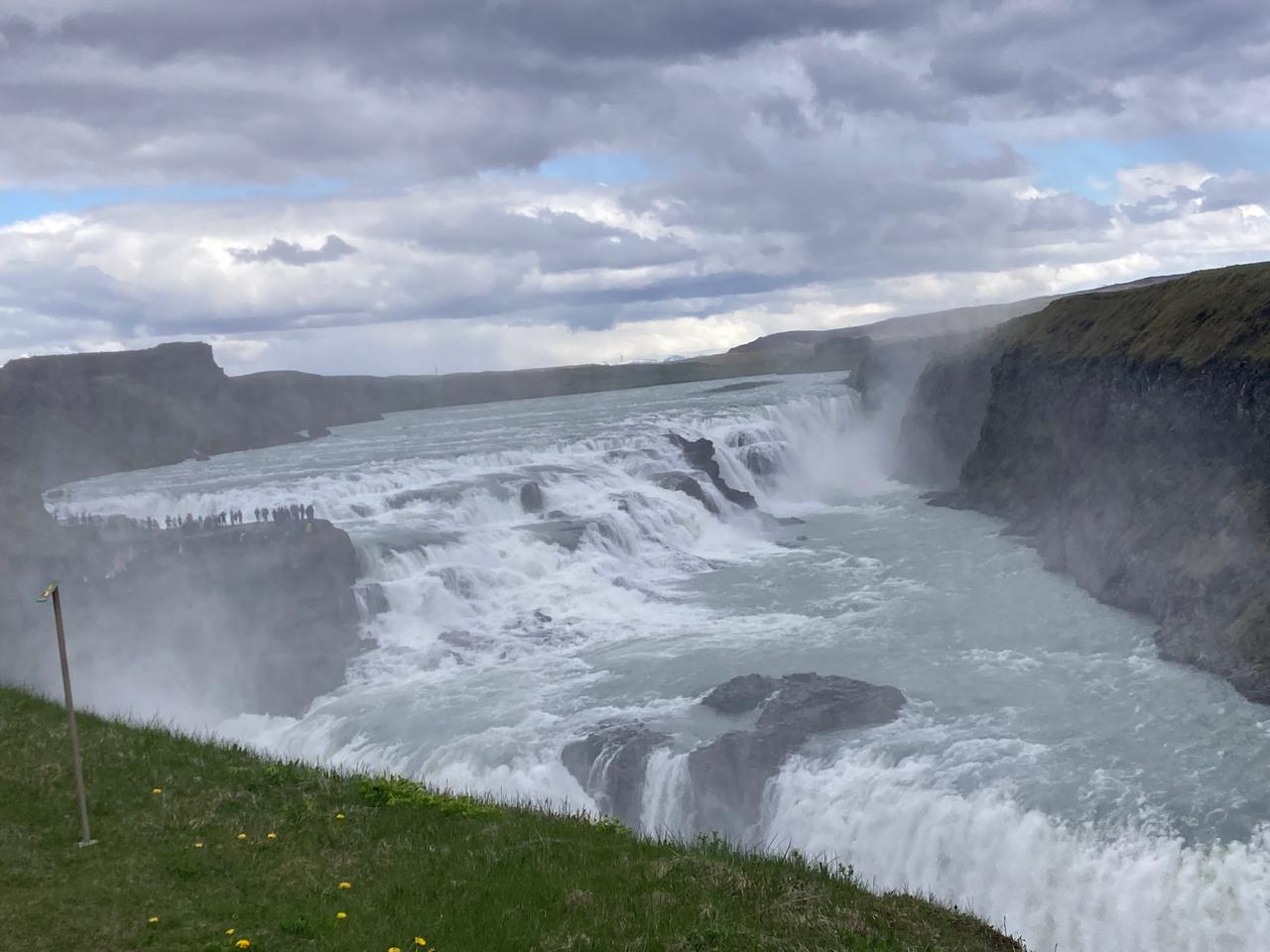 A wide waterfall churns over rocky cliffs under a cloudy sky, with visitors watching from a nearby overlook