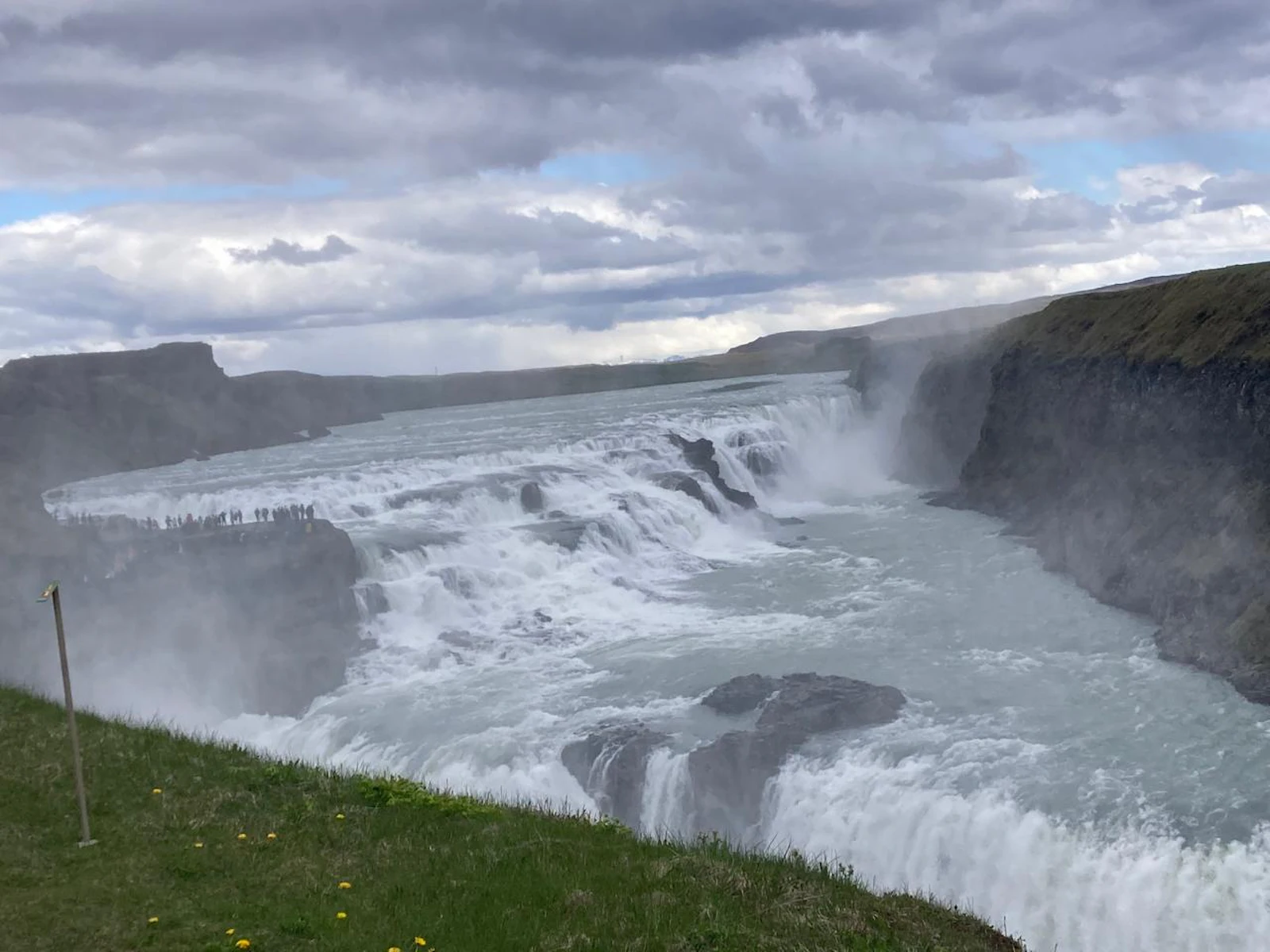 A wide river plunges over a two-step waterfall into a misty canyon below a grassy bank