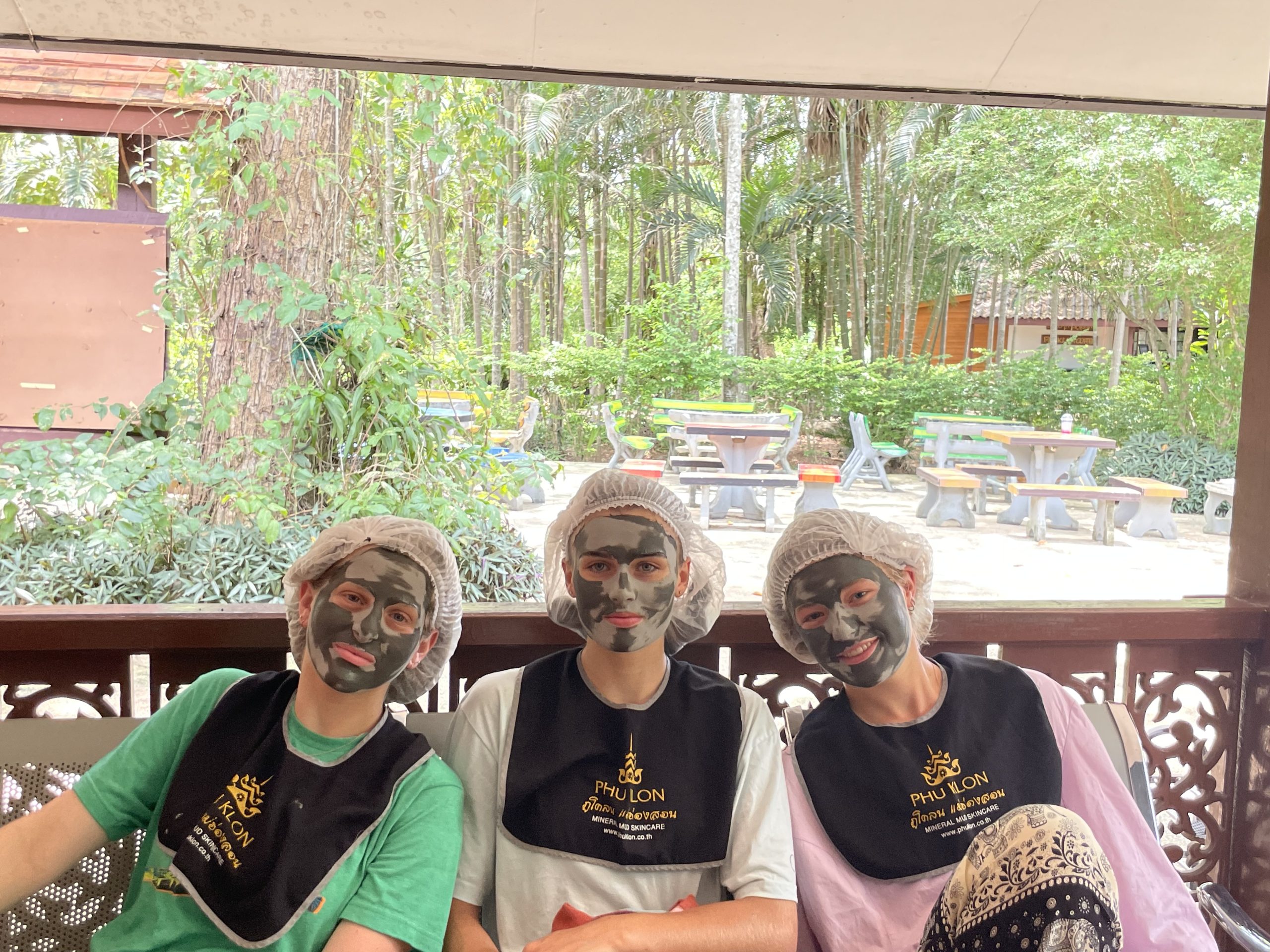 3 girls sitting down with a mud face mask on, outside on a bench