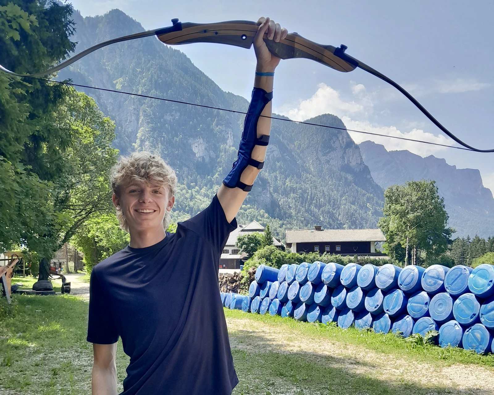 A teen traveler holds a recurve bow above his head, with forested mountains behind