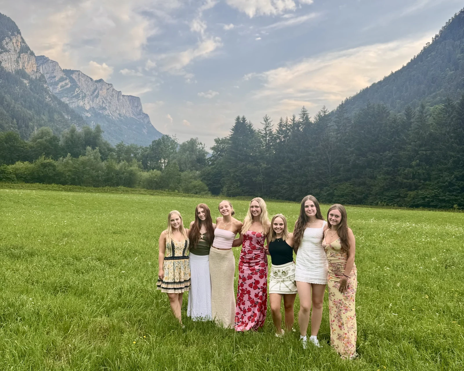 A group of teen travelers in summer dresses stands in a valley meadow below forested mountains at dusk